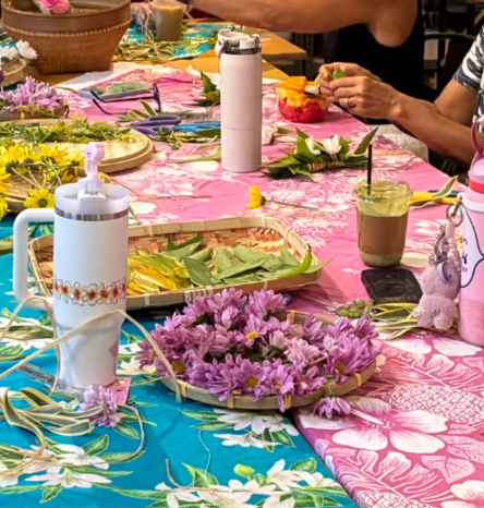 A table decorated with a pink and blue floral tablecloth, surrounded by people preparing flowers and arranging flower bouquets, with bottles, cups, and other supplies visible.