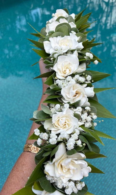 A person's arm with a gold watch holding a cascading white flower arrangement with roses and baby's breath over a blue swimming pool.