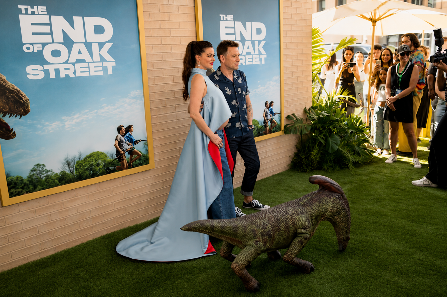 People attending a promotional event for 'The End of Oak Street' film, standing next to movie posters on a brick wall, with a plastic dinosaur on artificial grass and a crowd taking photos and videos in the background.