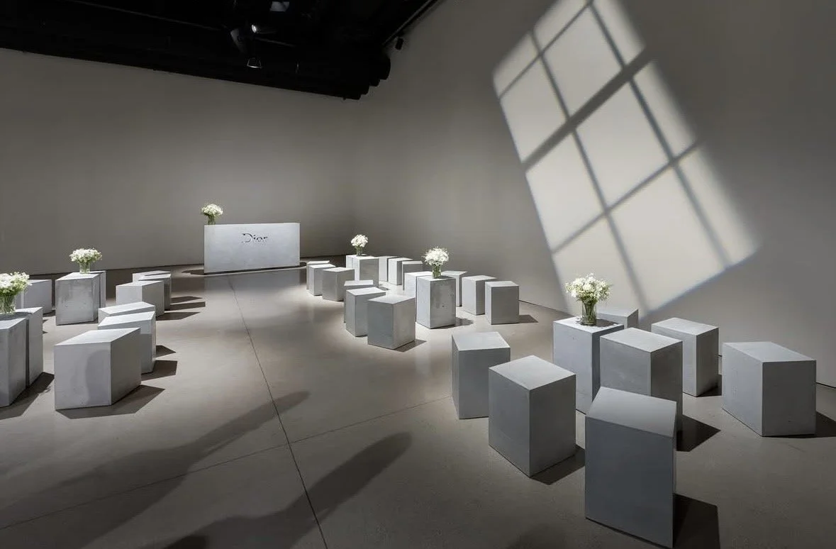 An art installation featuring rows of white pedestals with white flowers in vases, lit by sunlight and casting shadows through a window at a Dior event.