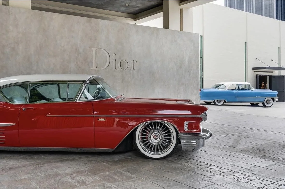 Vintage cars parked outside Dior store with a beige wall and modern building in the background.