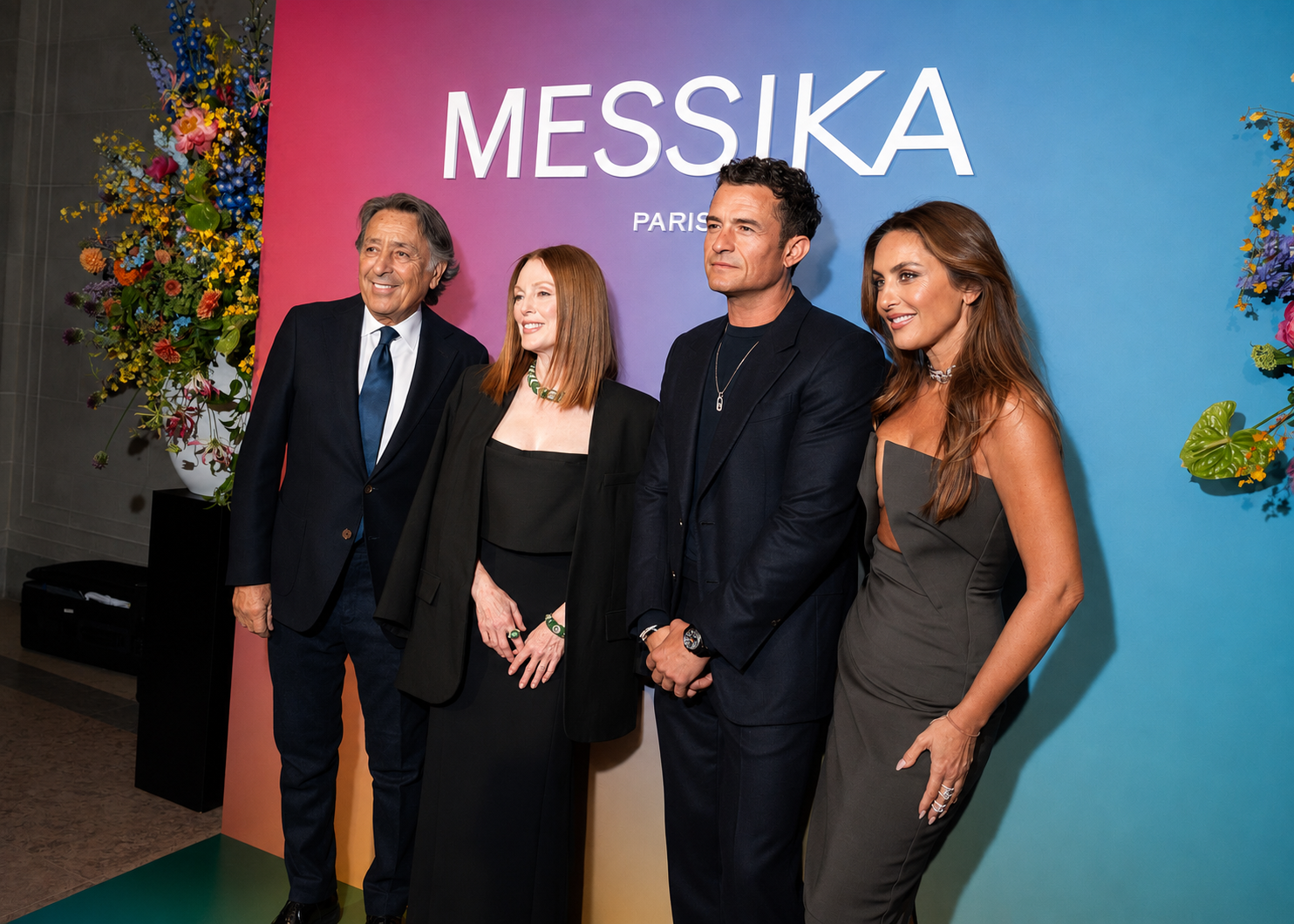 Four people standing in front of a colorful backdrop with the text 'MESSIKA PARIS'. They are dressed in formal attire, smiling, with flowers on either side of the backdrop.