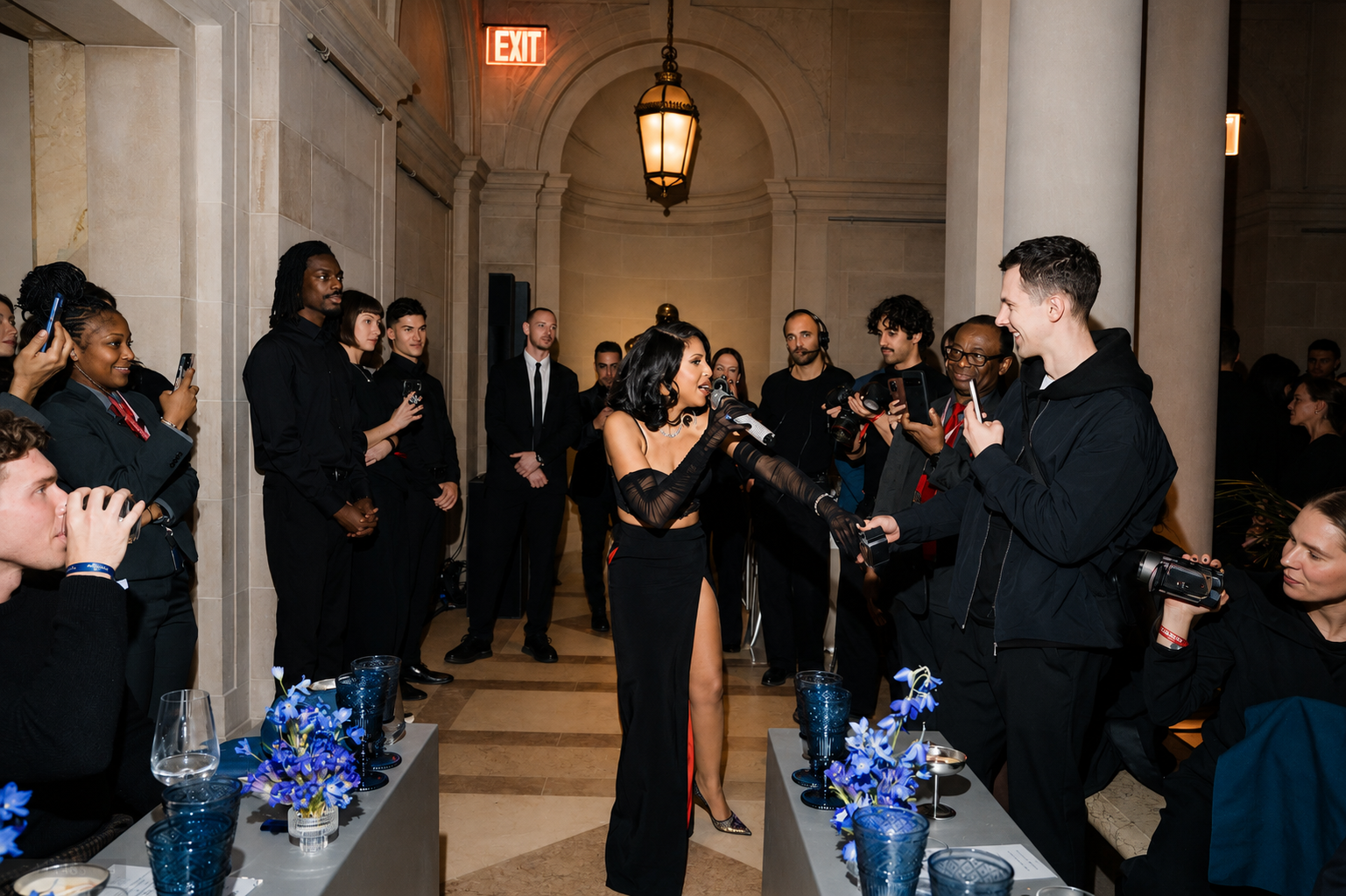 A woman in a black evening gown with a high slit is speaking into a microphone while being interviewed by journalists with cameras and microphones. The event takes place in an elegant indoor setting with a high ceiling and columns, with several onlookers and photographers observing. The women are dressed formally, and there are flower arrangements on the tables in the foreground.
