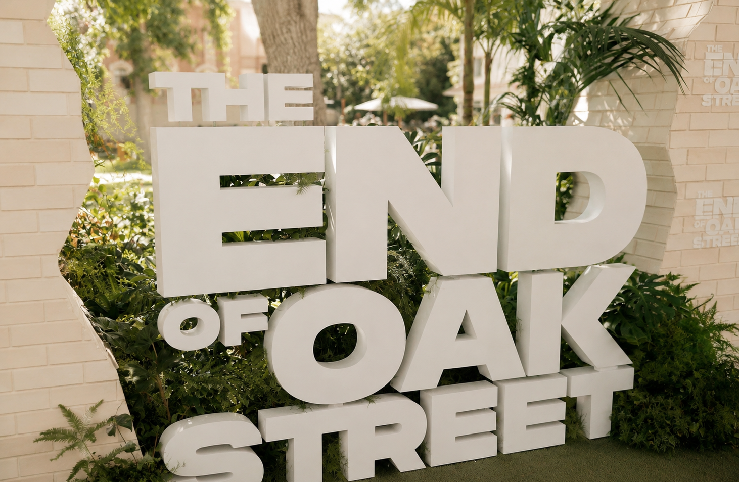 Large white block letters spell out "THE END OF OAK STREET" amid greenery on a sidewalk, with trees and buildings in the background.