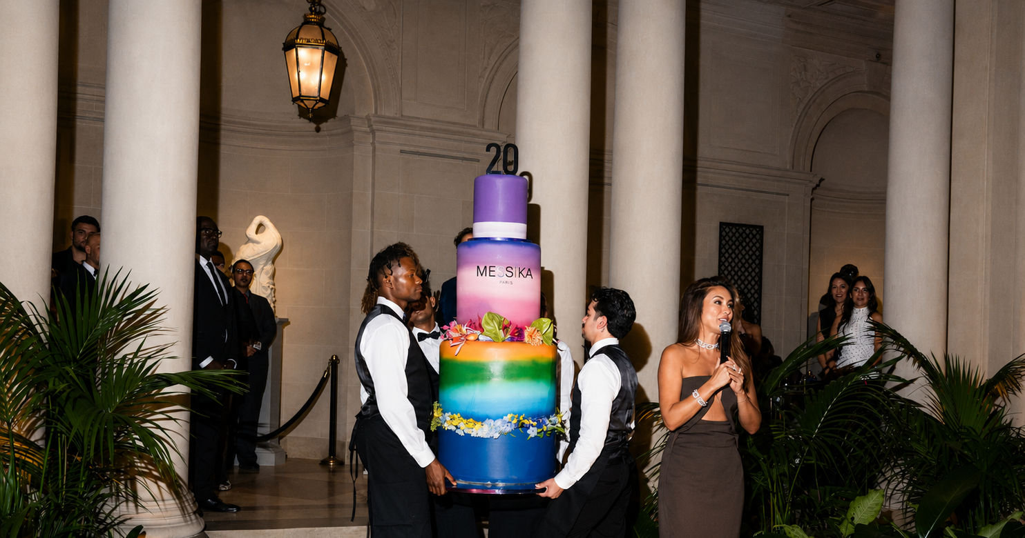 People holding a large, colorful birthday cake with a purple top, rainbow layers, and floral decorations inside a grand hall with tall columns, greenery, and a woman speaking into a microphone.