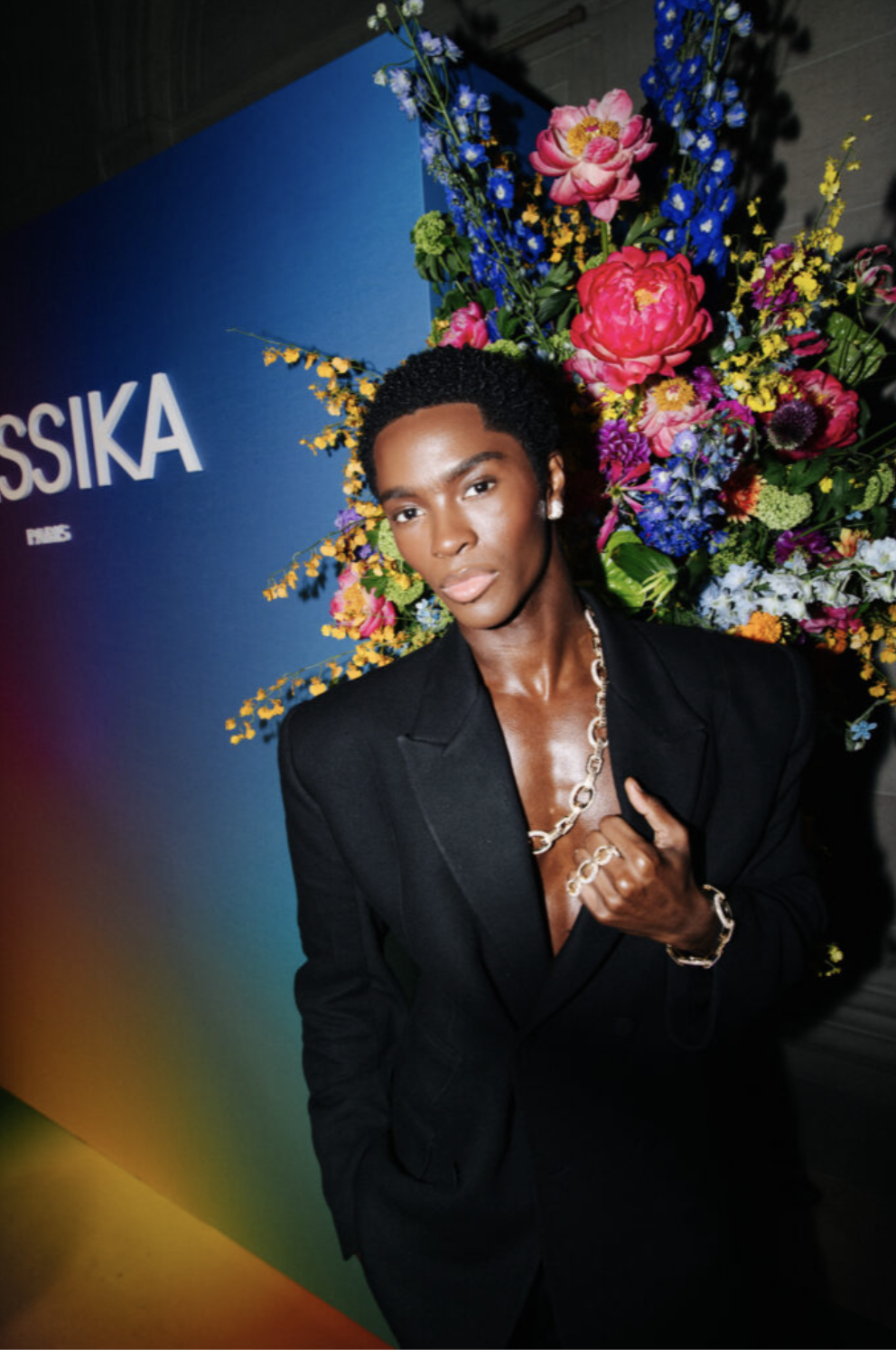 A person with dark skin and short curly hair wearing a black blazer, a chunky silver necklace, and a bracelet, posing in front of a colorful floral backdrop at an event.