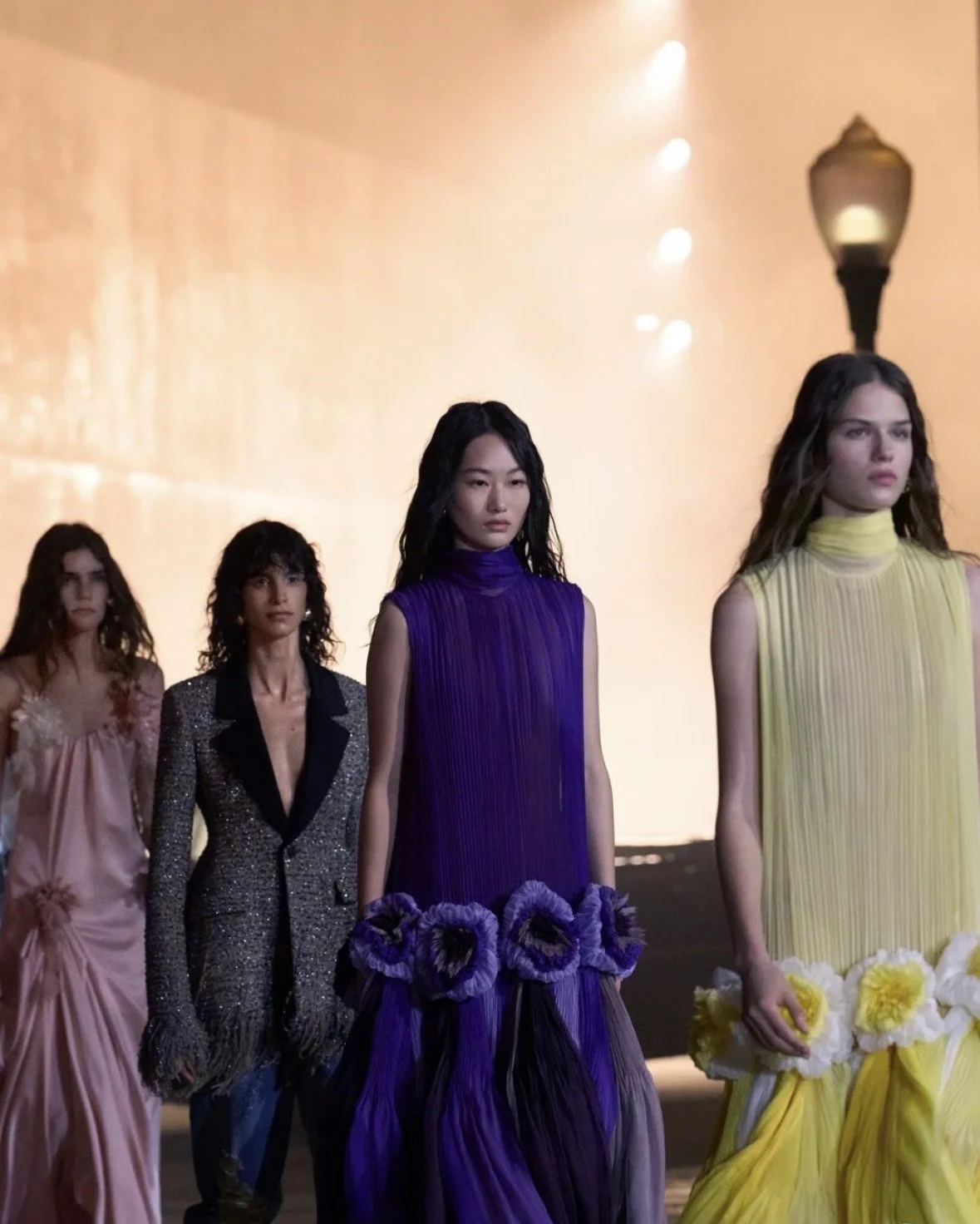 Four models walking on a runway, wearing colorful evening gowns, with a background of an orange sky and a lamp post.