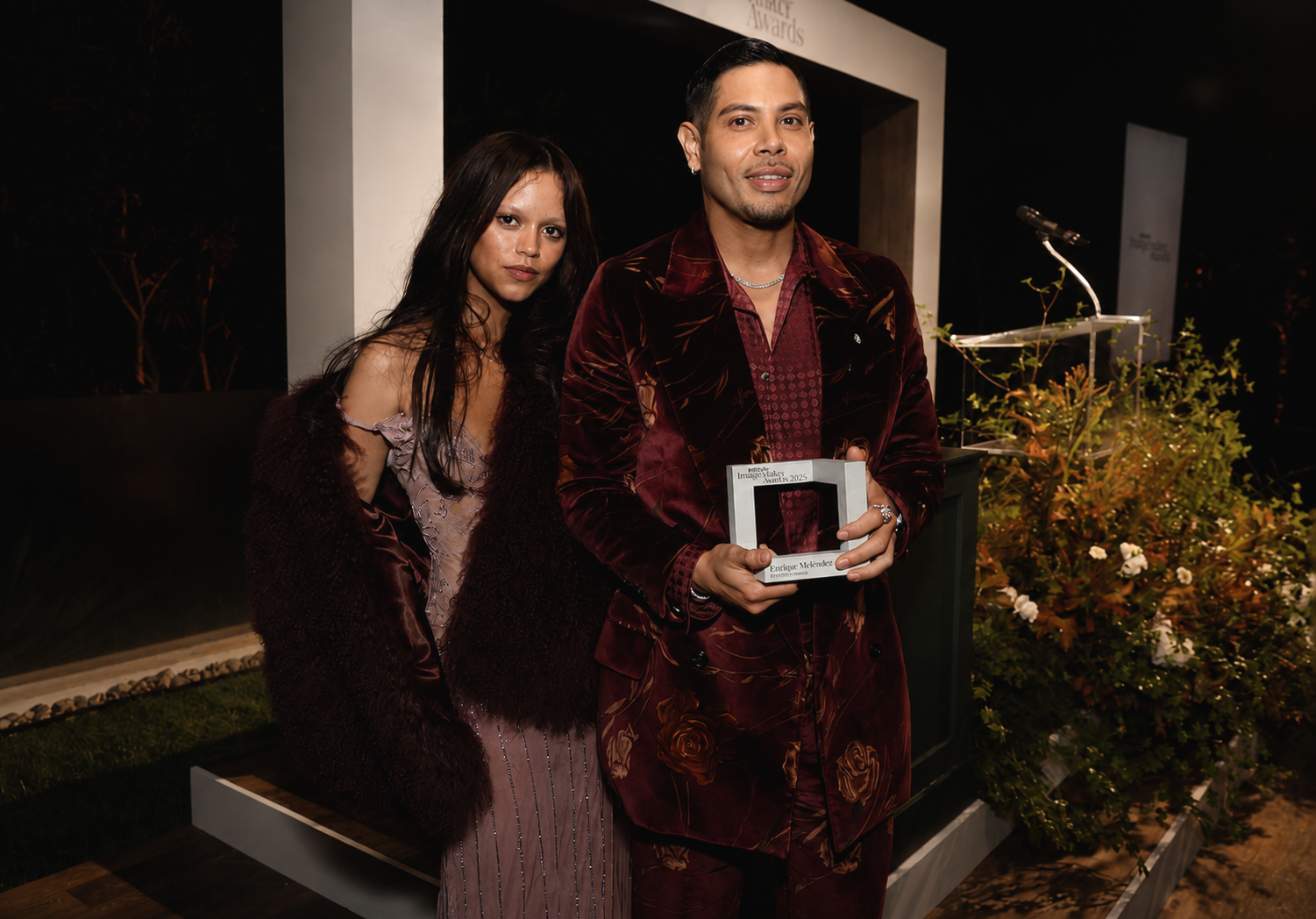 A man and woman dressed in formal attire standing together on a stage, with the man holding an award plaque. The man is wearing a velvet jacket with floral embroidery, and the woman is wearing a shiny dress with a fur coat. There are plants and a podium with a microphone in the background.