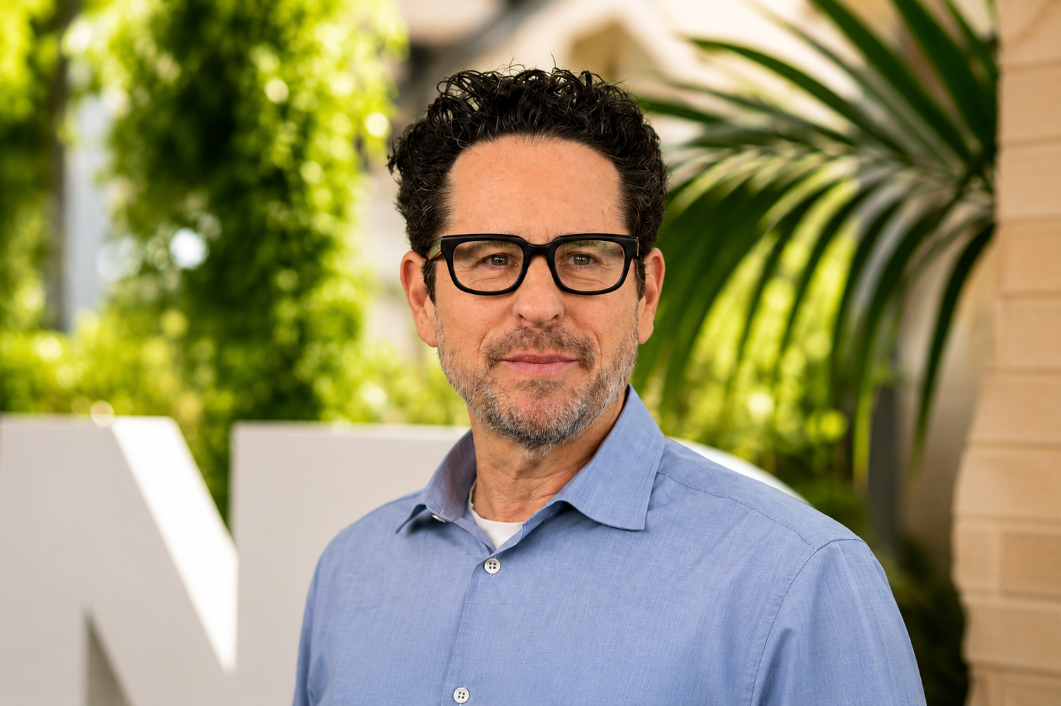 A man with dark, curly hair, wearing glasses and a blue button-up shirt, standing outdoors with a background of green foliage and a building.