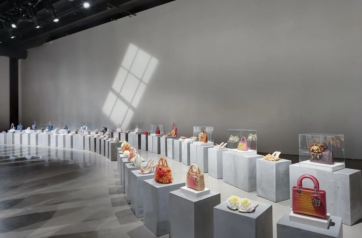 Display of handbags and shoes in an art gallery style on white pedestals with glass cases, illuminated by natural light through a large window reflected on the wall.