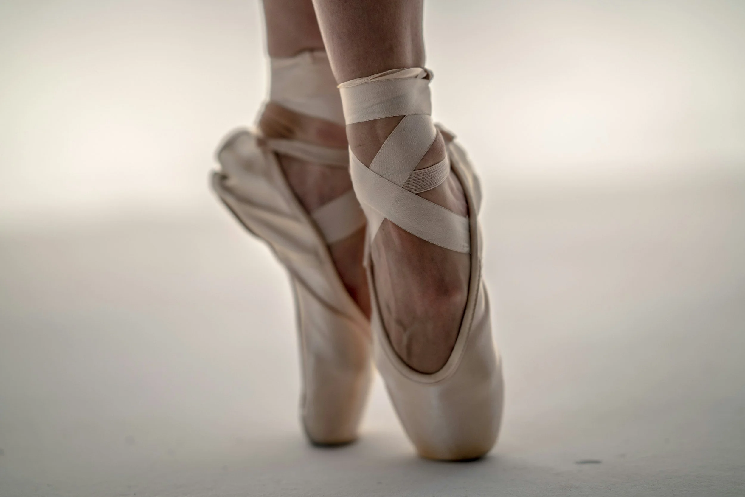 Close-up of a ballet dancer's feet in pointe shoes, wrapped with ribbons, standing on a white surface with a plain background.