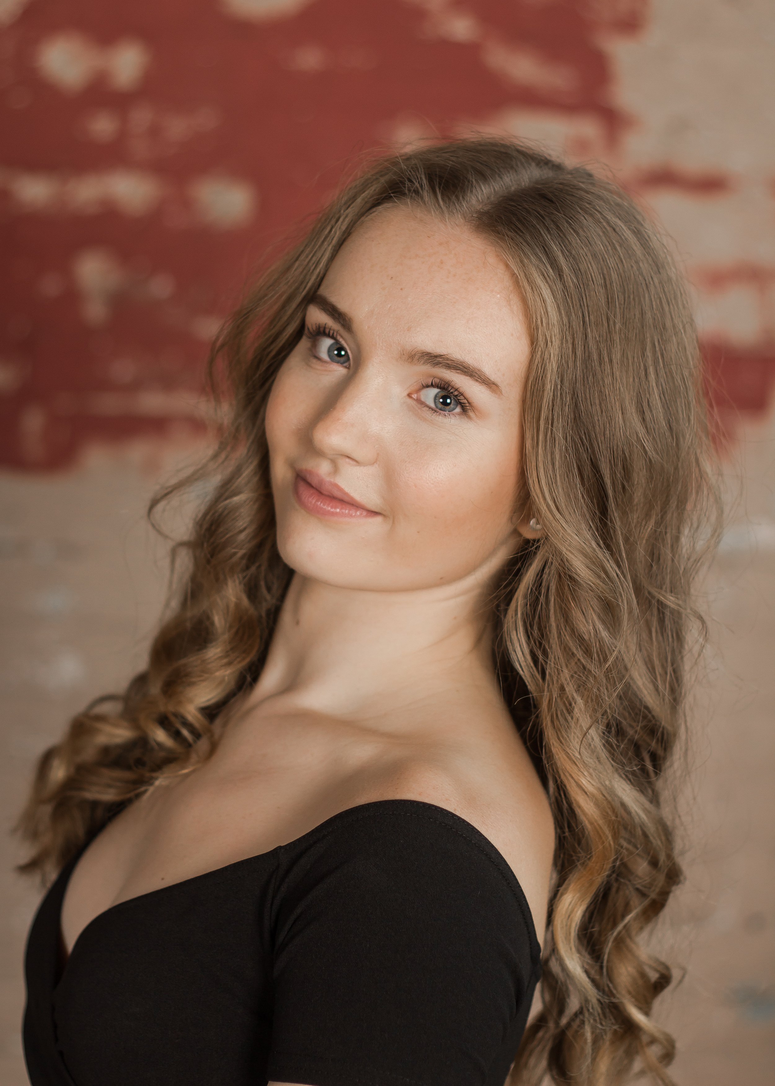 A young woman with long wavy hair, light skin, and blue eyes, wearing a black off-shoulder top, standing against a red and beige patterned background.