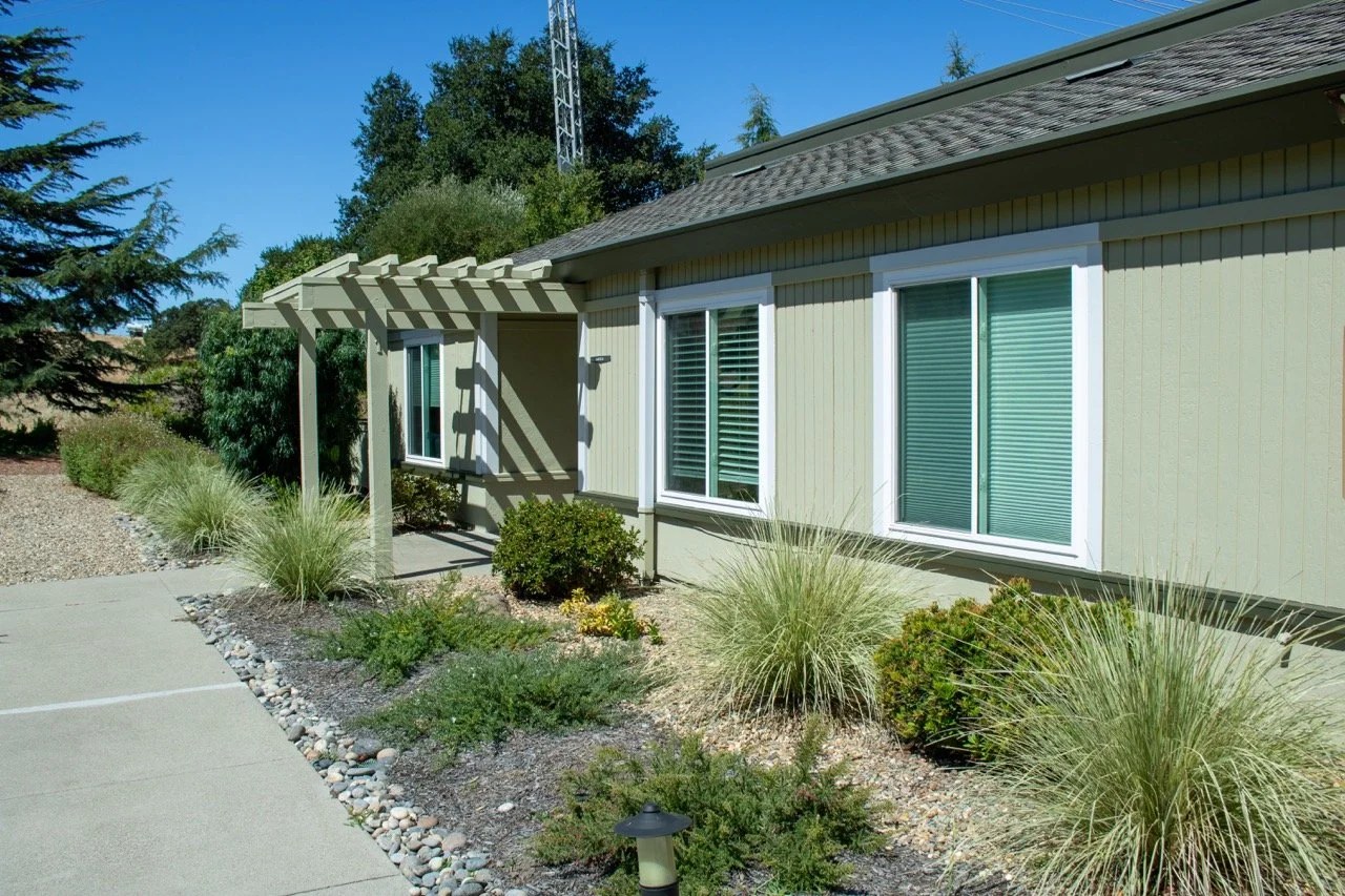 Exterior view of a house with a light green siding, white window frames, and a small covered patio with a pergola. Landscaping includes bushes, ornamental grasses, and a paved sidewalk.