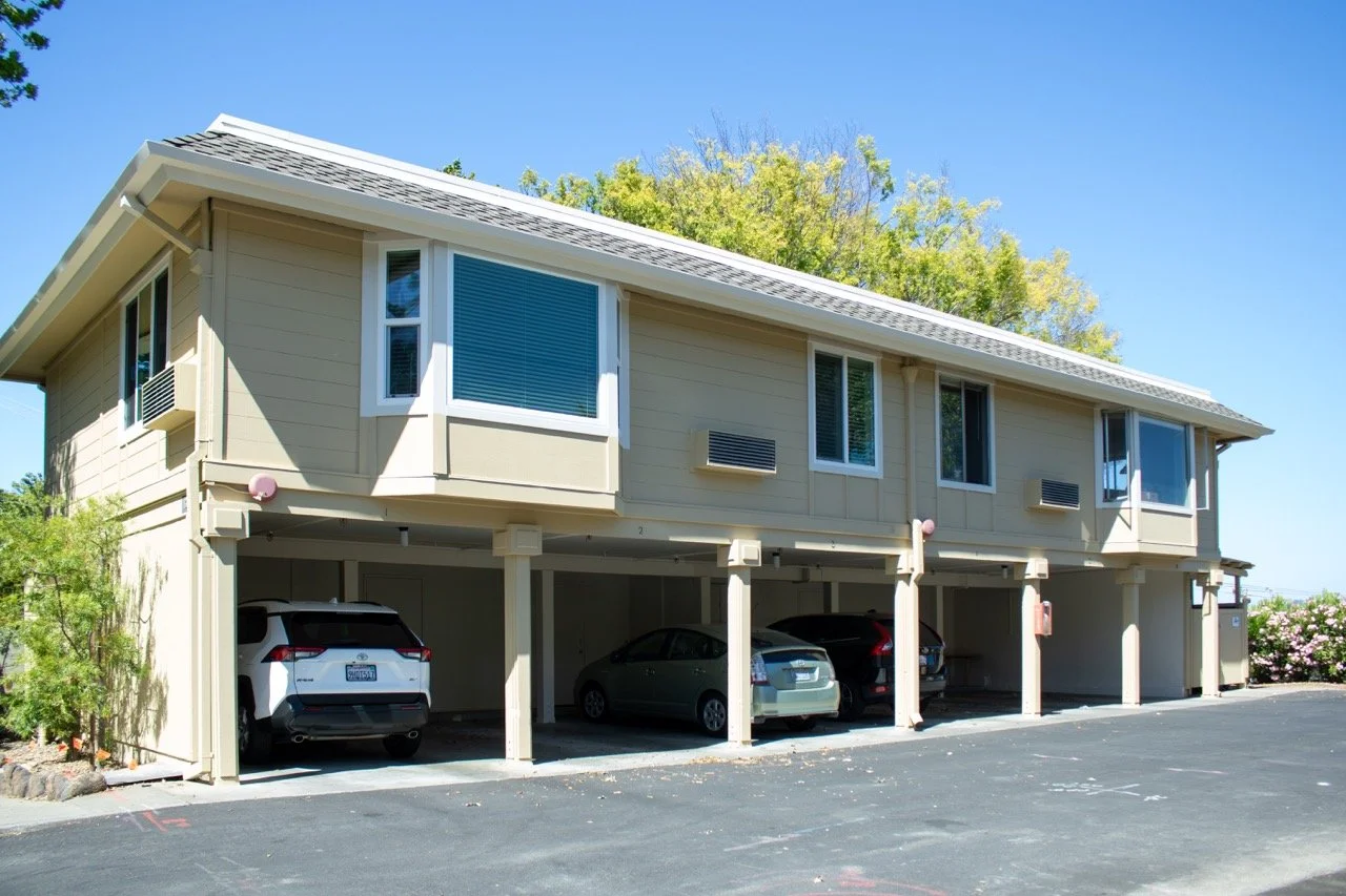 Multi-level residential building with parking on the ground floor, beige exterior, several windows and trees in the background, under clear blue sky.
