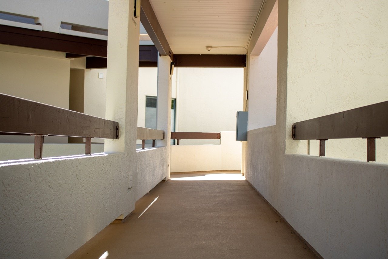 Empty outdoor balcony with beige walls, brown railings, and a concrete floor, illuminated by sunlight.