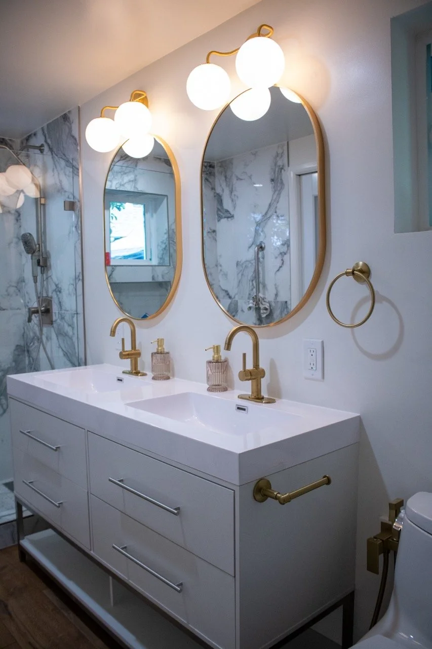 Modern bathroom vanity with double sinks, white countertop, gold fixtures, oval mirrors, and wall-mounted light fixtures with spherical bulbs.