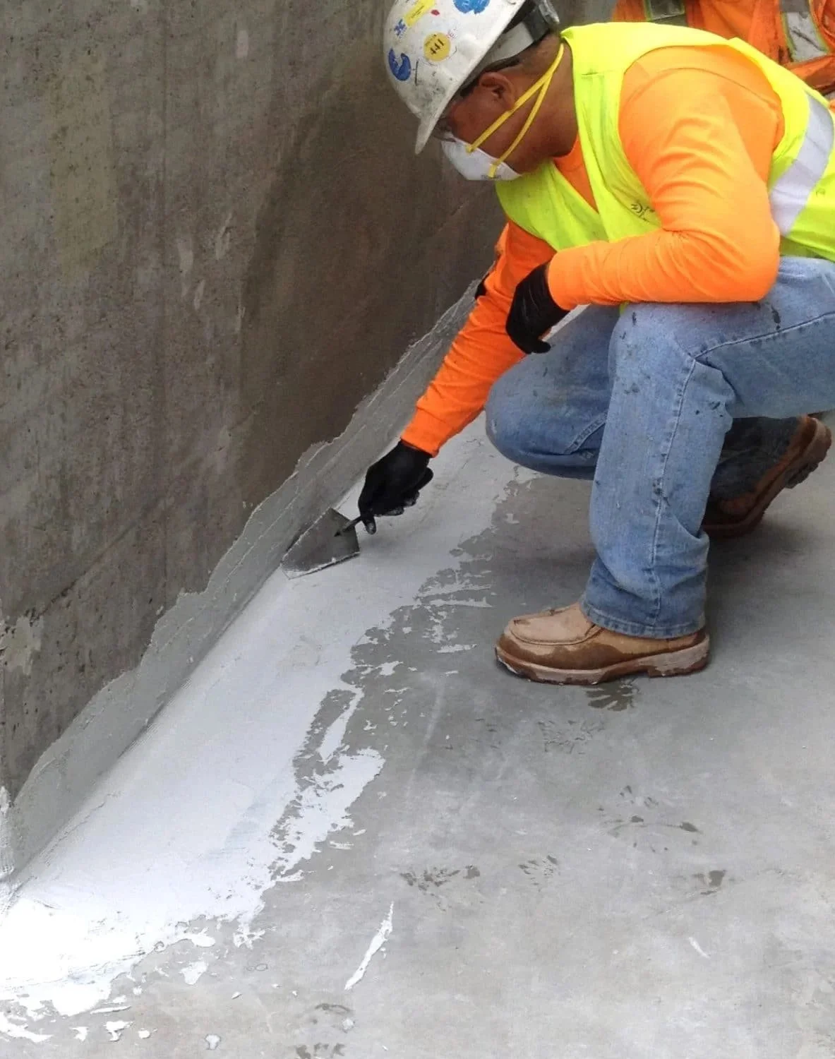 Construction site showing foundation waterproofing with black membrane, drainage pipe, and surrounding excavation.