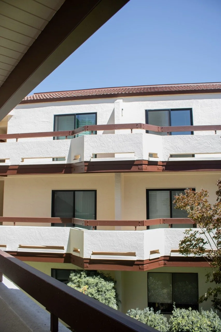 View of a multi-story apartment building with white walls, black-framed windows, and brown railings on the balconies, set against a clear blue sky.