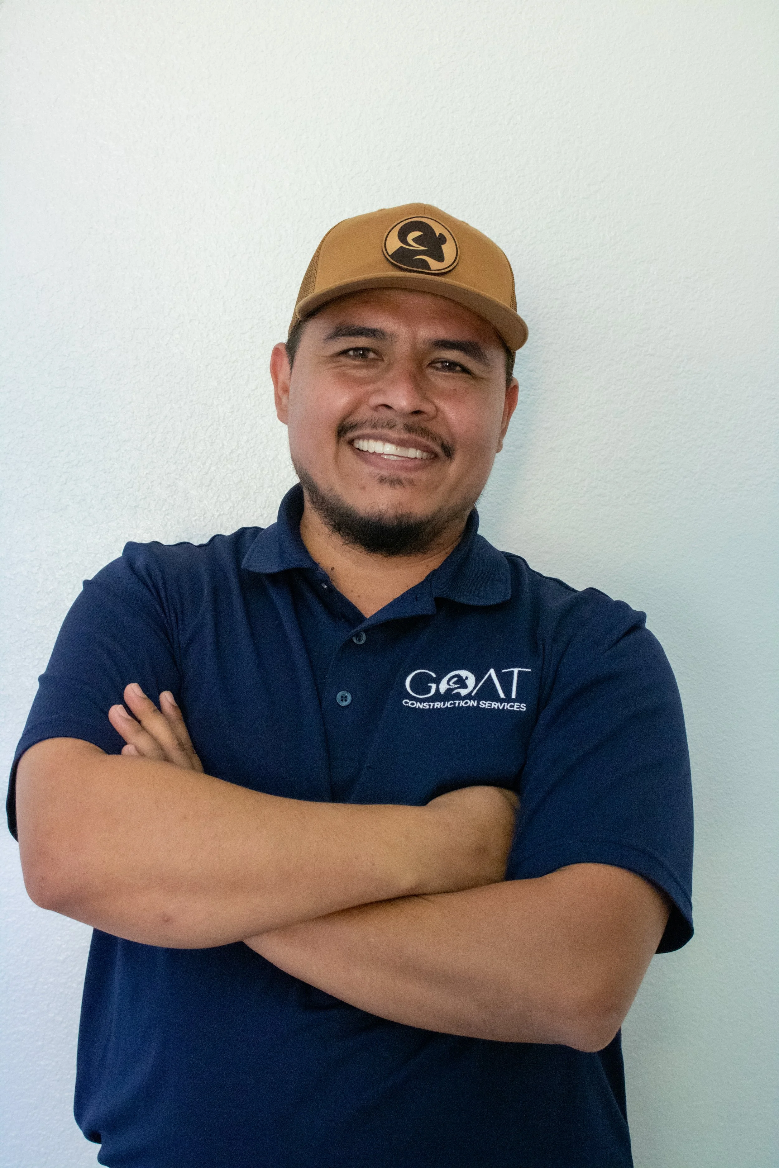 A man in a navy blue polo shirt with the logo of GOAT Construction Services, standing against a white wall, smiling with arms crossed, wearing a tan baseball cap with a logo on the front.