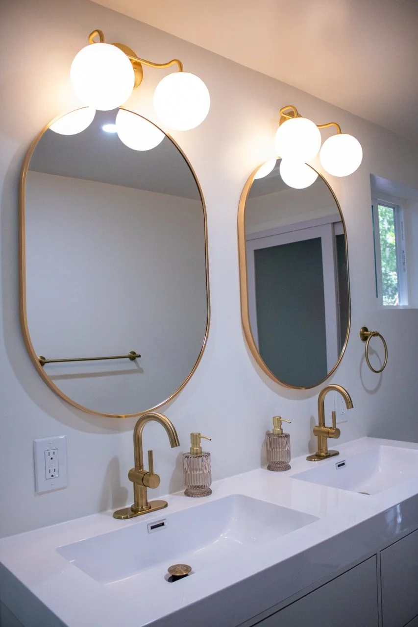 Bathroom with two white sinks, gold faucets, two large oval mirrors with gold frames, and three globe lights above each mirror. There is a small window on the right.