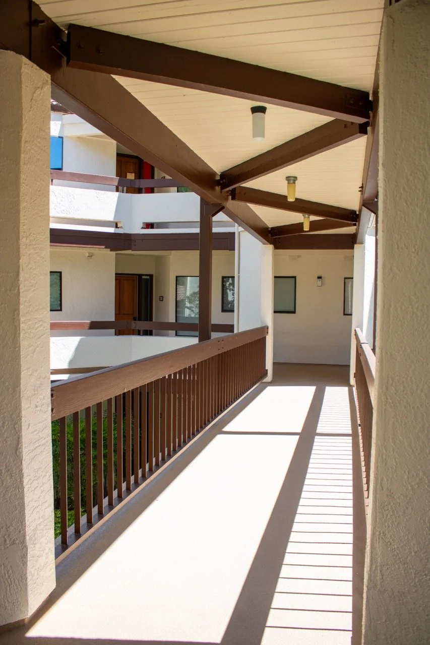 View of a corridor on an apartment balcony with wooden railings and overhangs, casting shadows on the concrete floor.