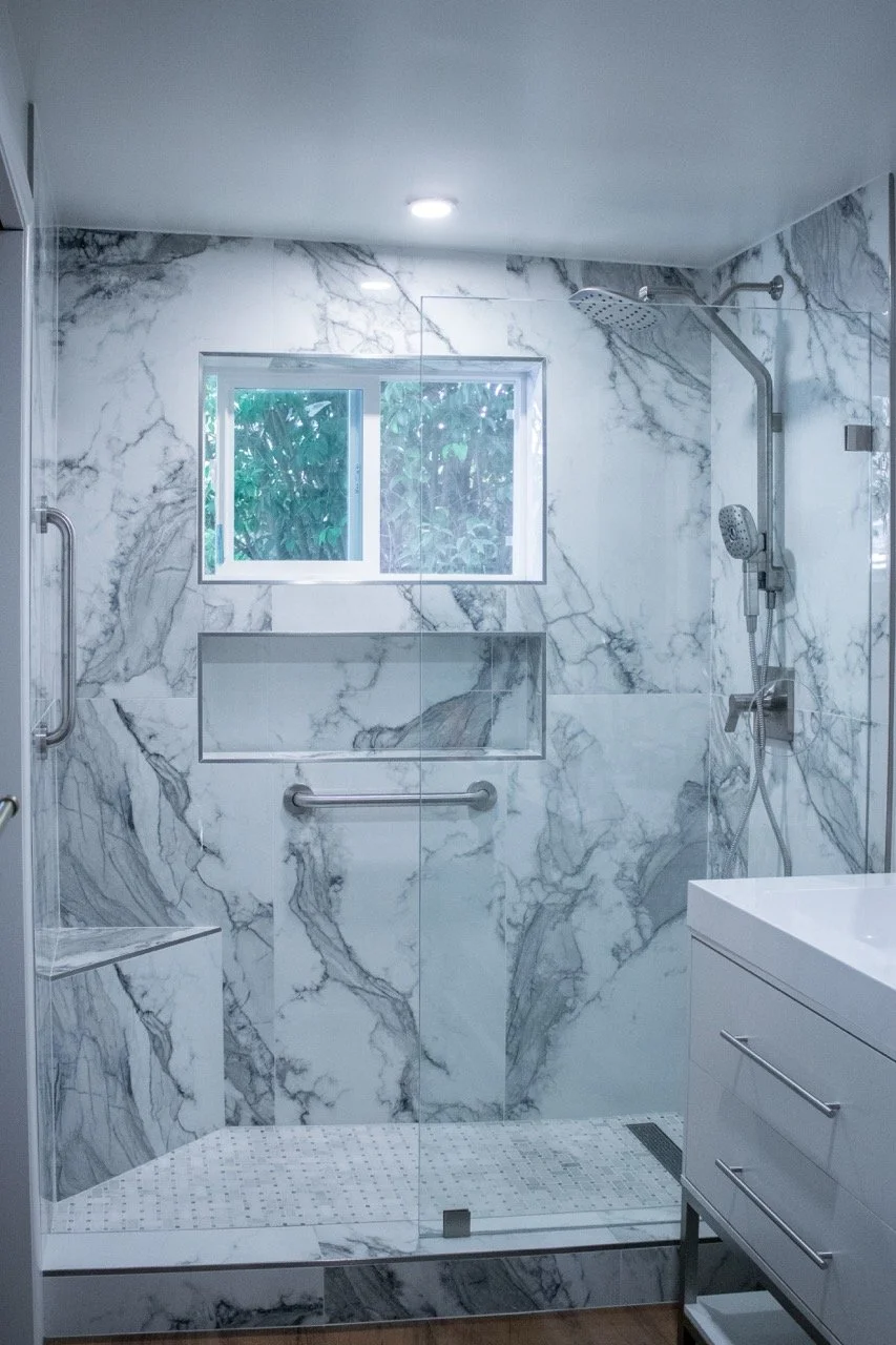 A modern walk-in shower with marble walls, a small window, a built-in bench, separate rain and handheld showerheads, and a glass enclosure.