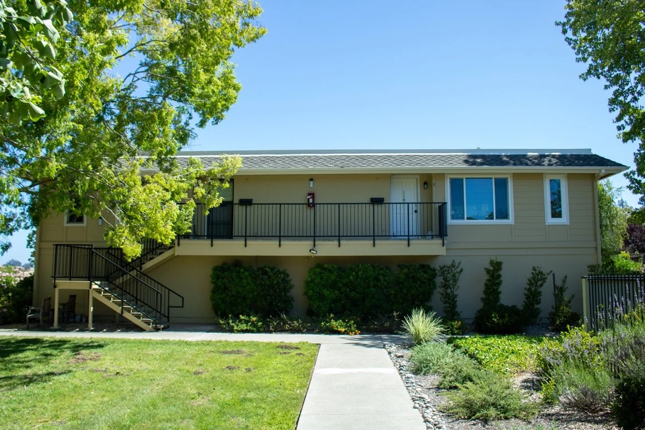 Exterior view of a beige, two-story residential building with a front balcony, outdoor stairs, and landscaped yard with trees and bushes on a sunny day.