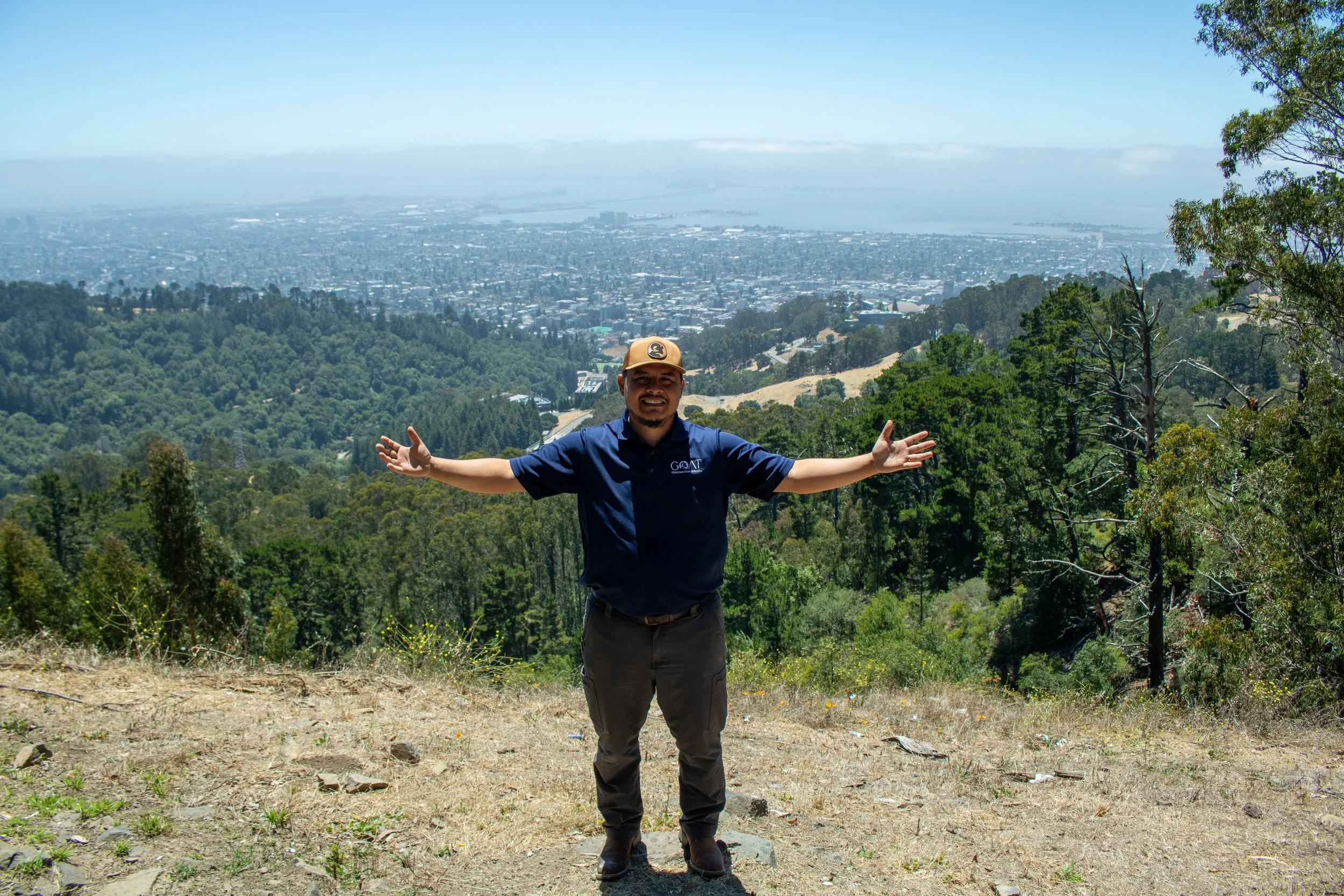 A man standing with arms open, smiling, on a hiking trail surrounded by trees, overlooking a city and water in the distance.
