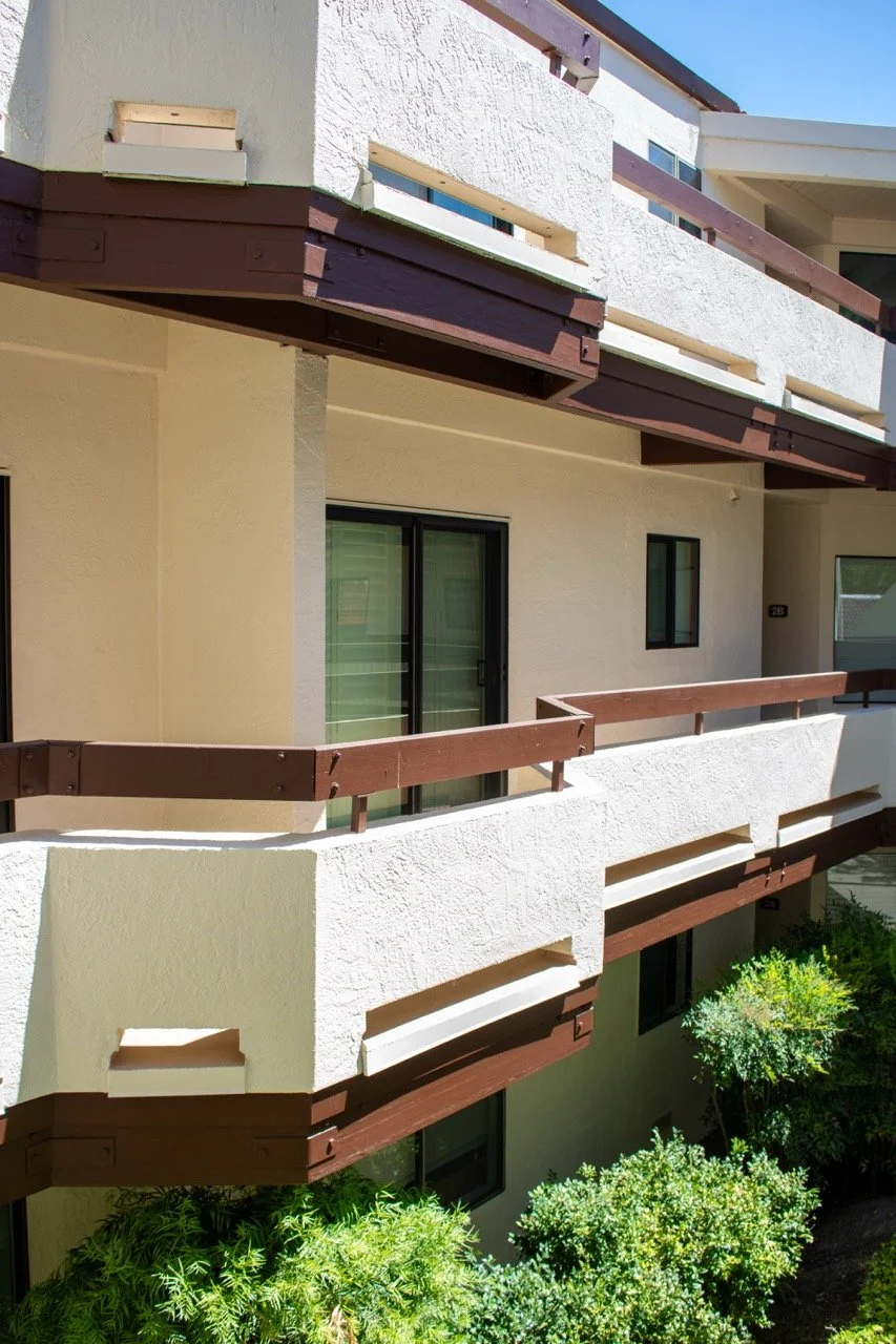 Balcony of a modern apartment building with white textured walls, wooden brown railings, and large windows, with green shrubs below.