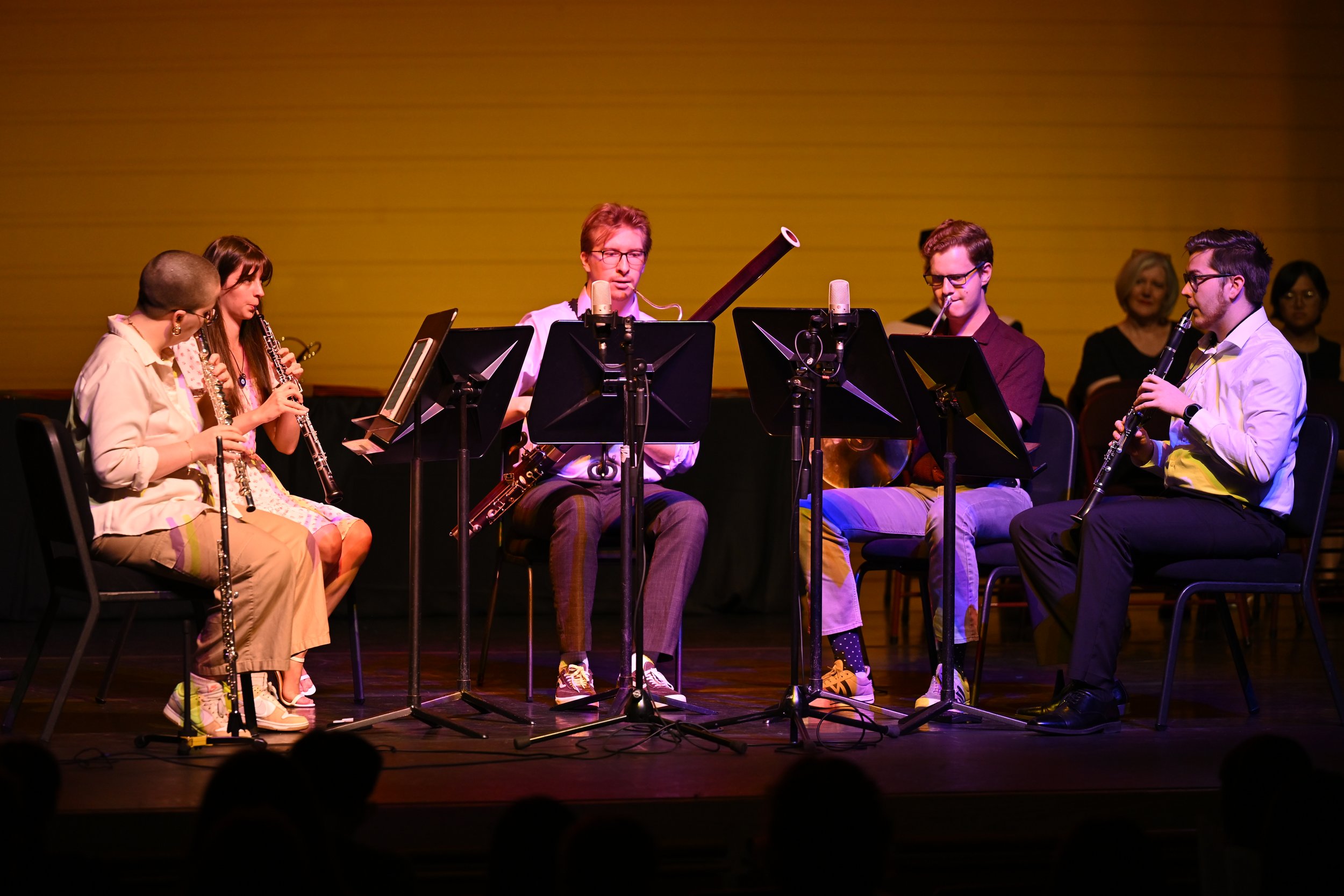 A group of five musicians playing wind instruments during a concert, seated on stage with music stands and microphones, with several people watching in the background.