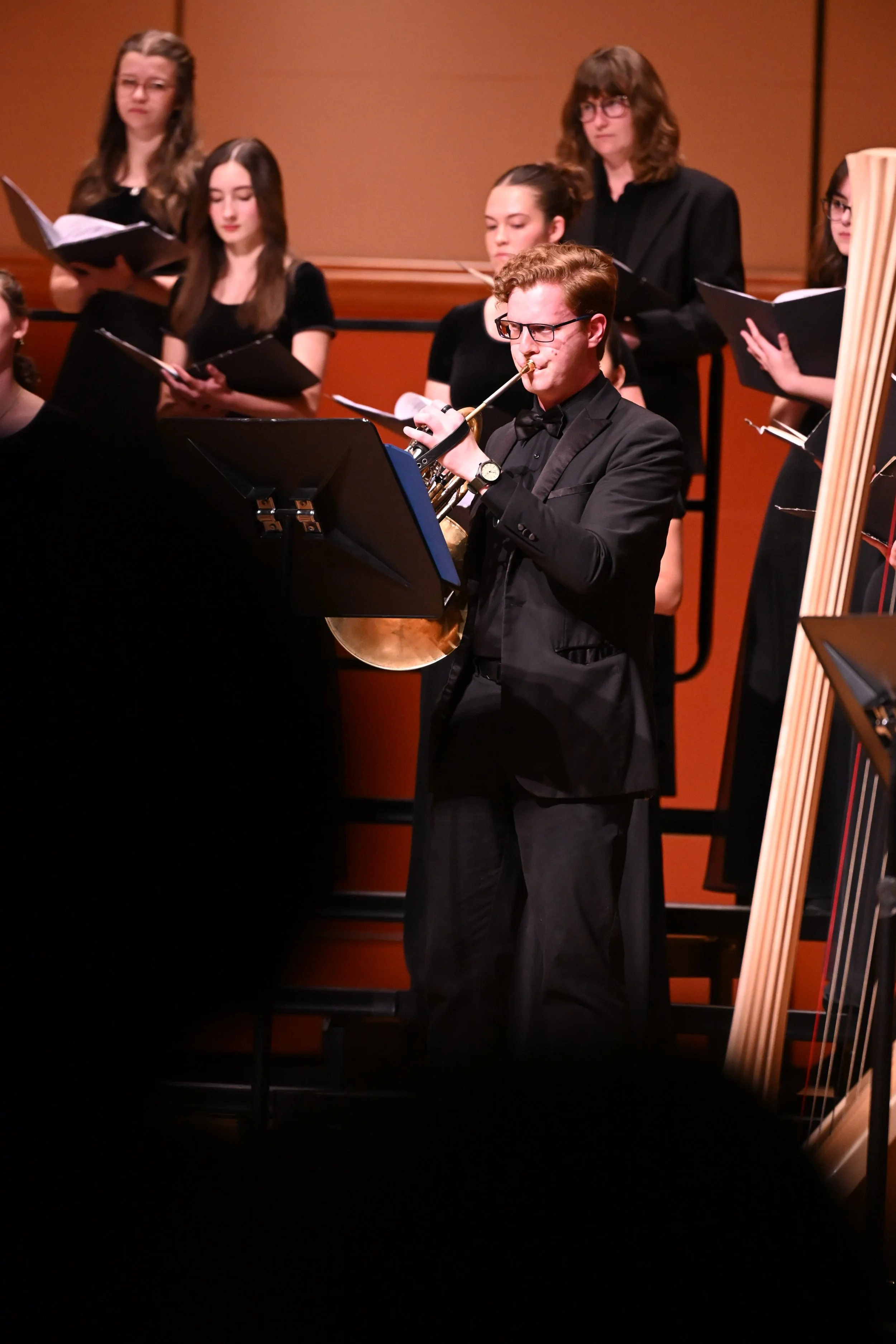 A man in a black tuxedo and glasses playing the trumpet during a choir performance, with several women standing behind him holding songbooks and dressed in black.