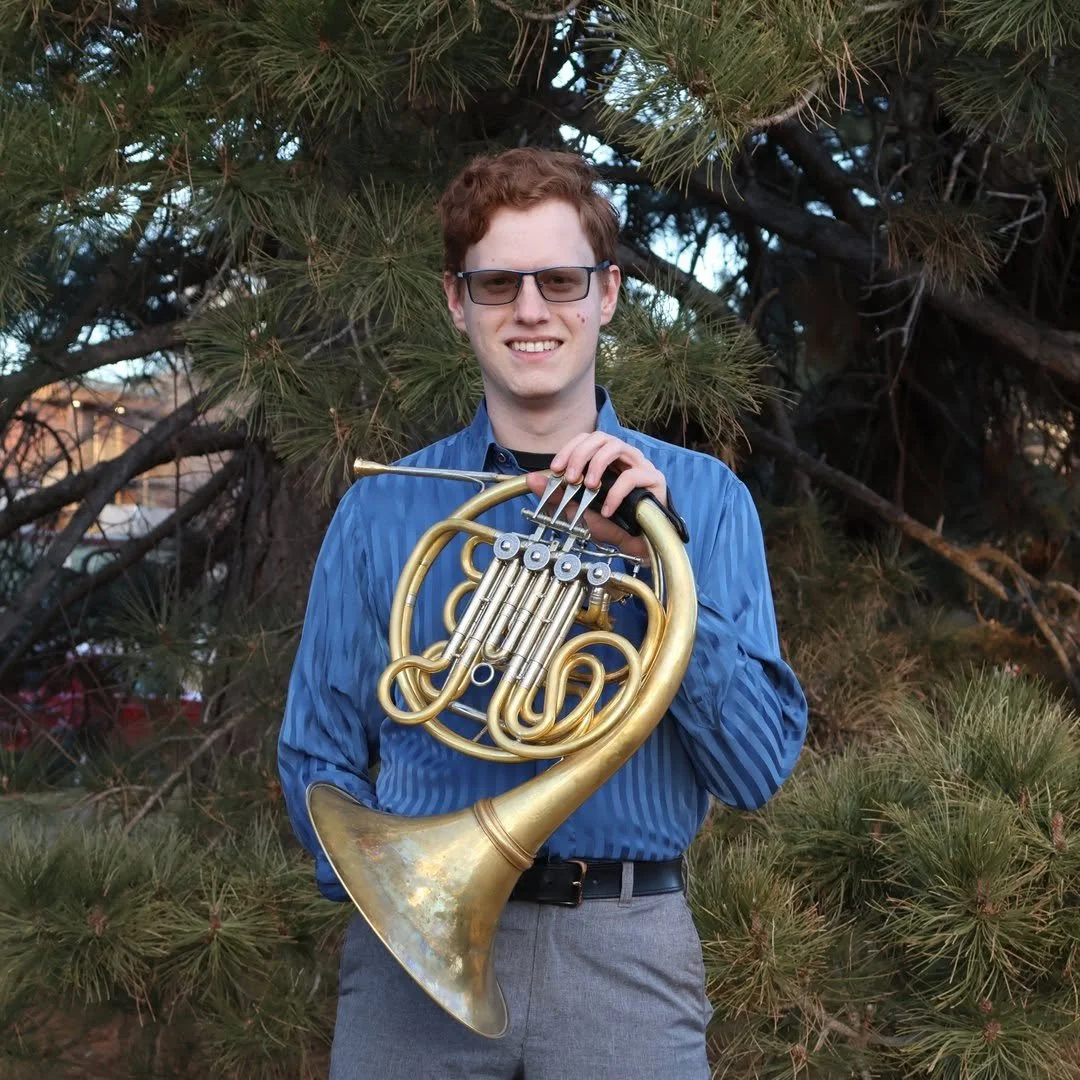 A young man with curly red hair, wearing glasses and a blue striped shirt, holding an old brass French horn. He is outdoors in front of a pine tree, smiling at the camera.