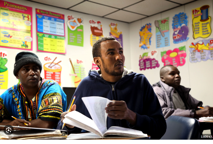 A classroom with three adult students sitting at desks, attentive, surrounded by colorful educational posters on the wall.