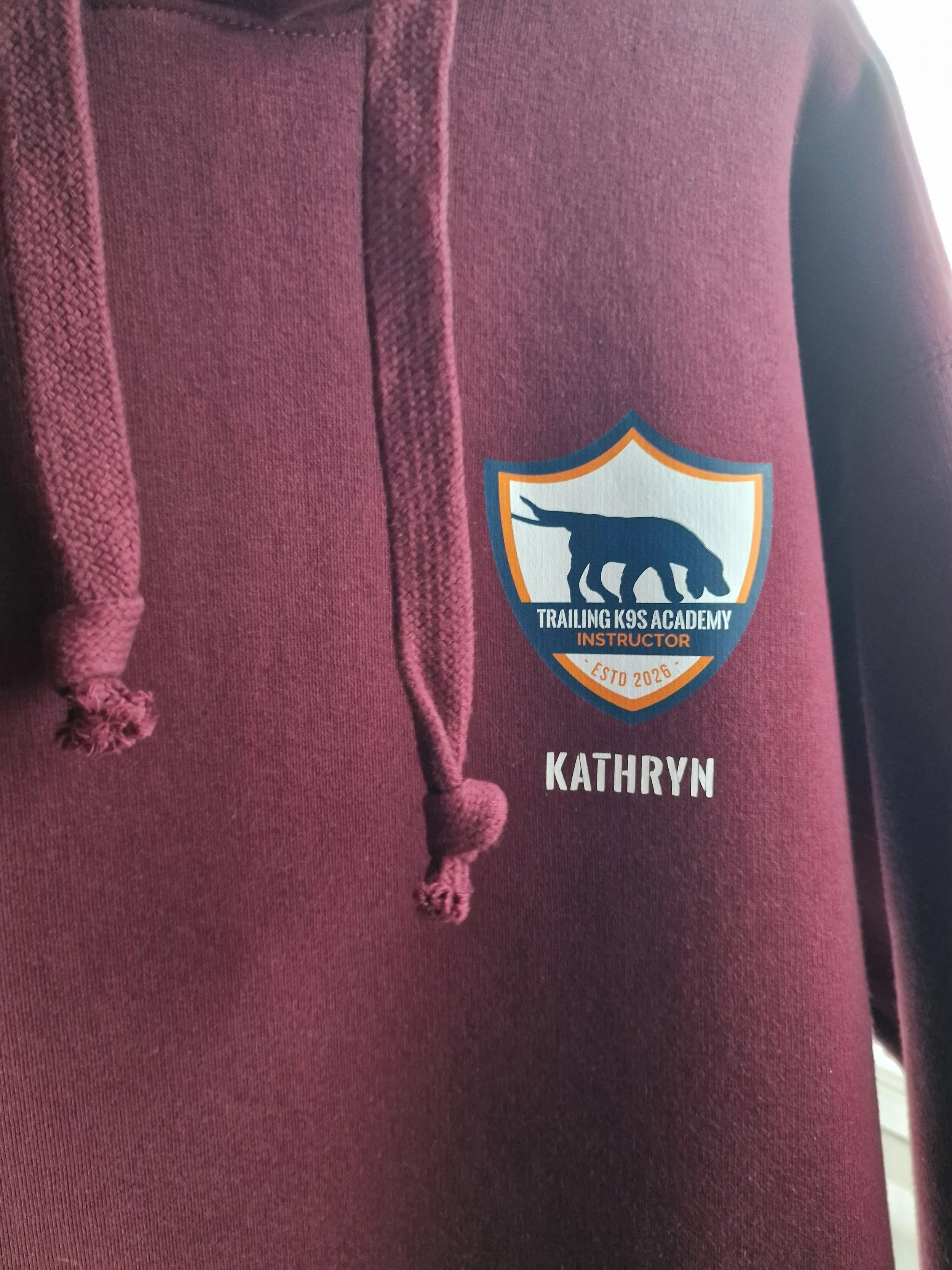 Close-up of a maroon hoodie with a trail running academy badge on the chest, displaying a bear silhouette, the text "TRAILING K9S ACADEMY," and the name "KATHRYN" underneath.
