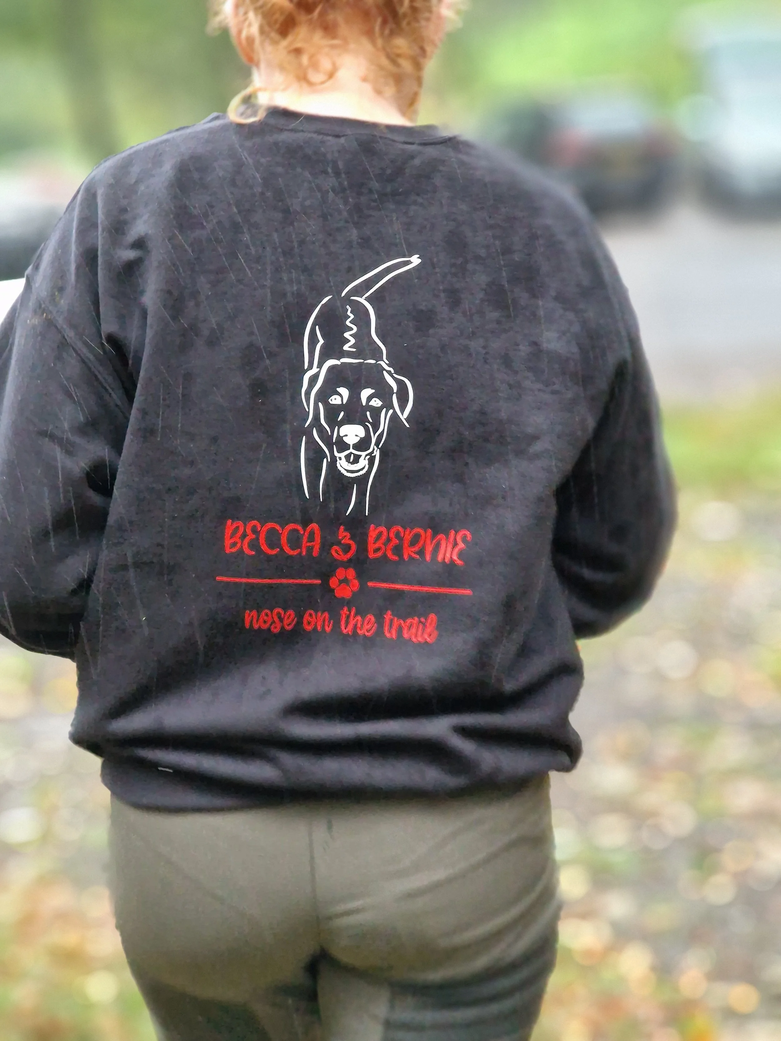 A person wearing a black hoodie with a white outline of a dog on the back and red text that reads 'BECCA & BERNIE' and 'nose on the trail'. They are outdoors with greenery and blurred vehicles in the background.