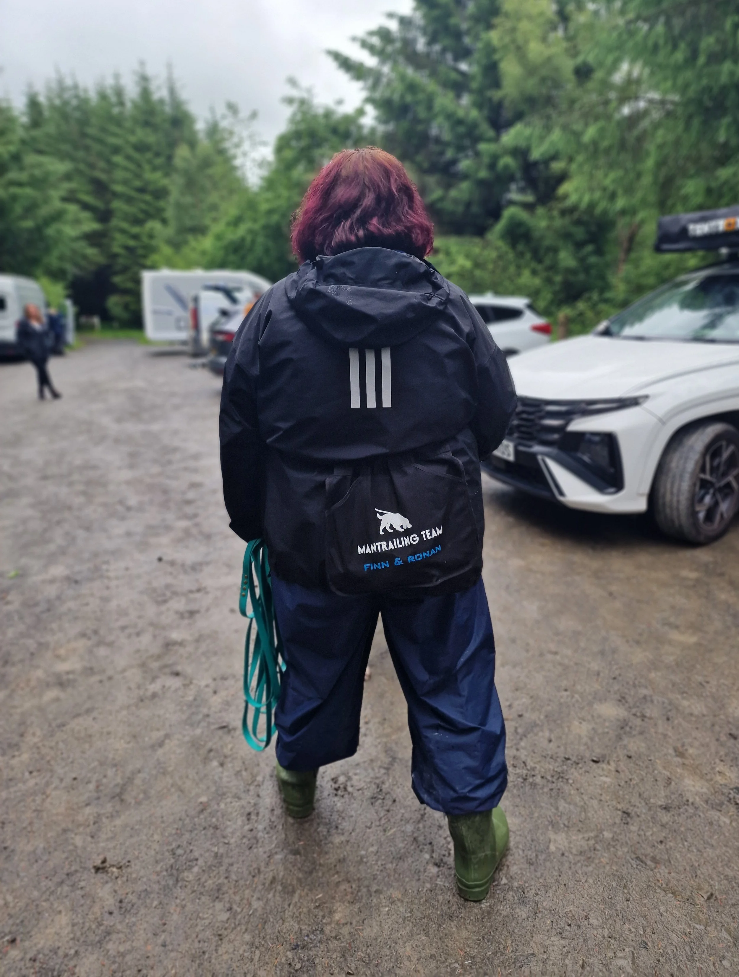 Person standing outdoors on a gravel surface, facing away, wearing a black jacket and Dog Training Apron with a logo, dark pants, and green rubber boots, holding climbing or safety ropes, with trees and parked cars visible in the background.