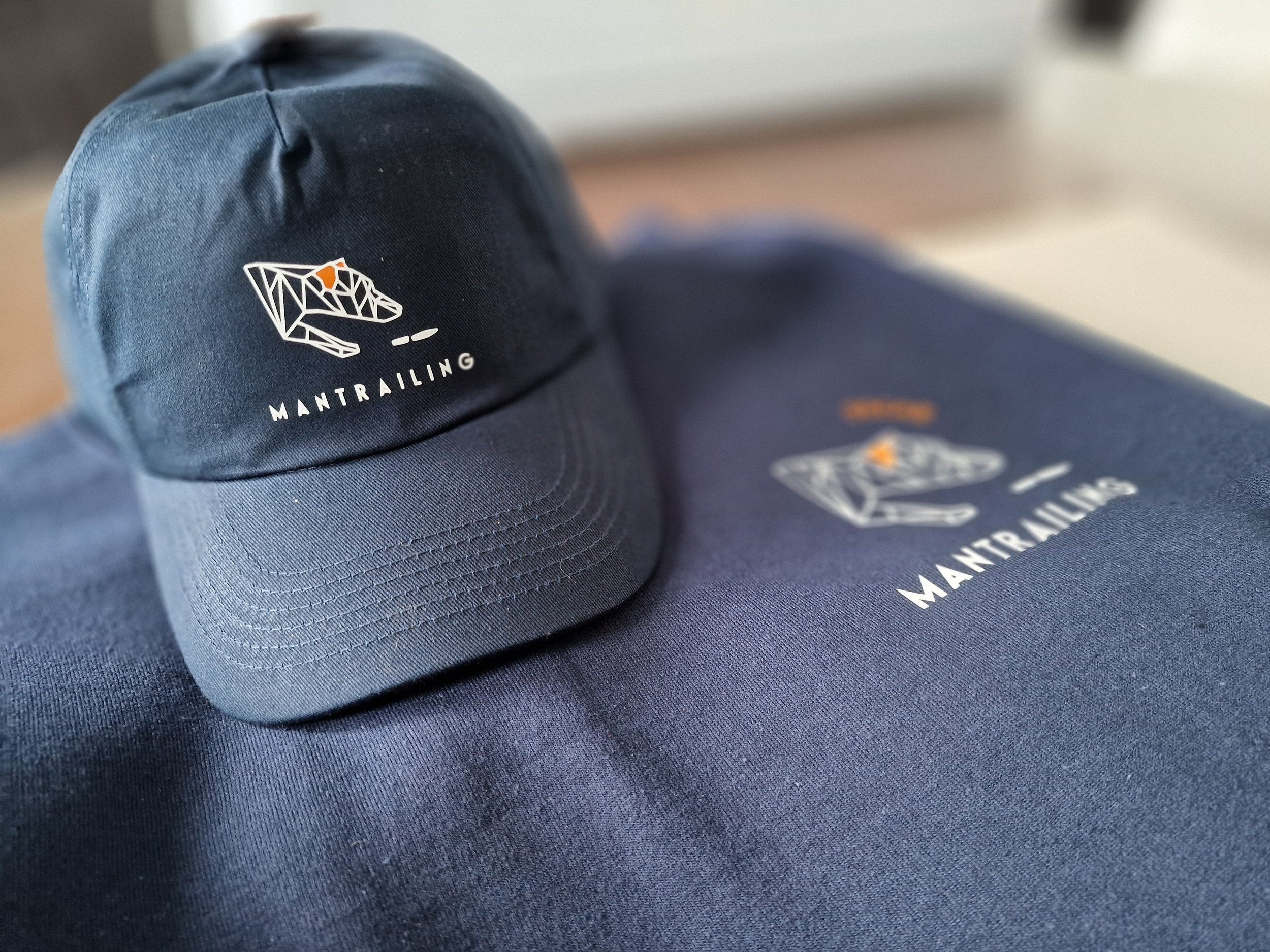 A navy blue cap with a geometric bear logo and the word "MANTRAILING" printed on it, resting on a navy blue fabric surface.