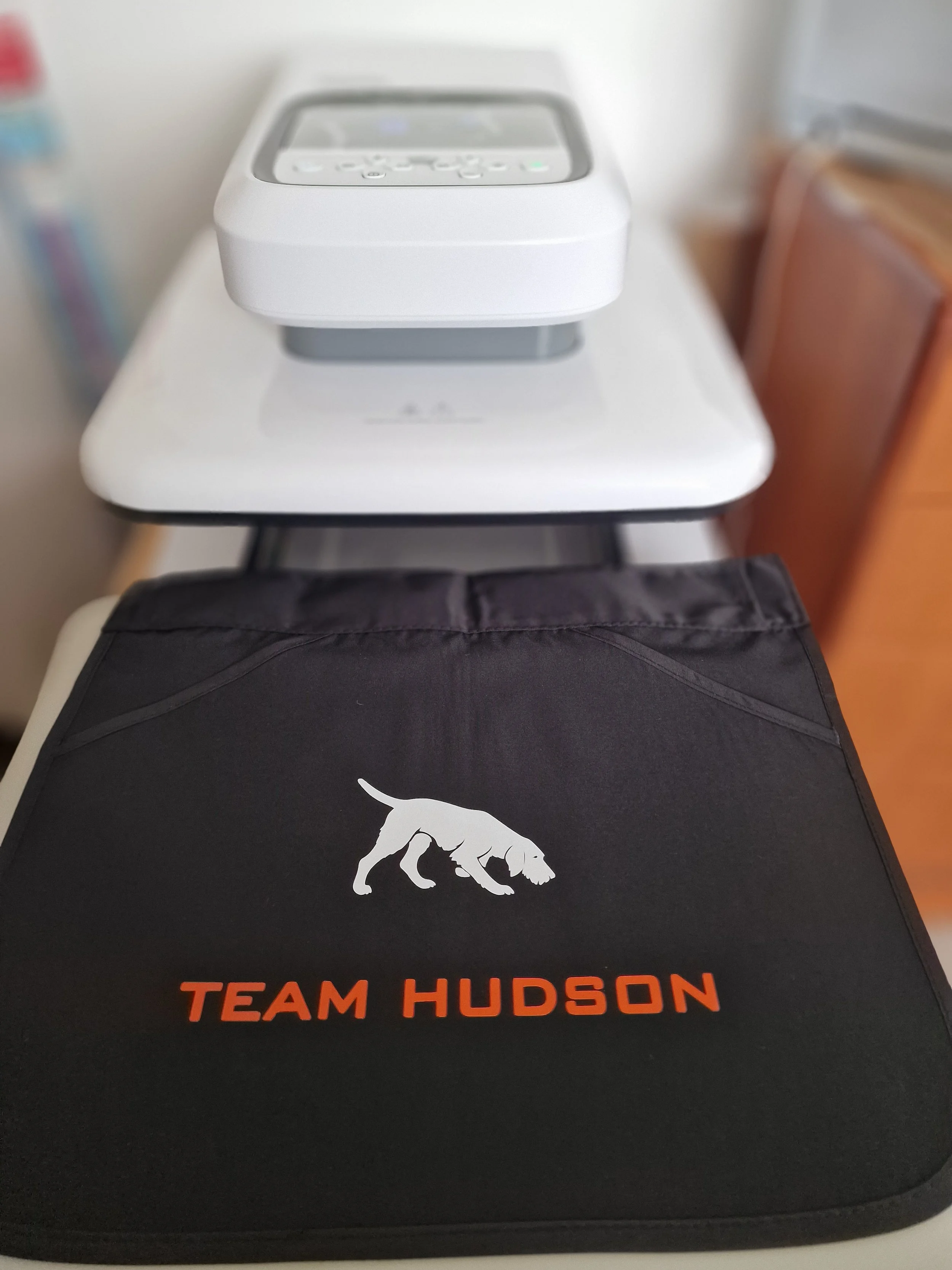 Close-up of a Dog Training Apron being heat pressed with an orange logo that reads 'TEAM HUDSON' and a white silhouette of a dog. On top of the heatpress, there is a white device with a screen and buttons.