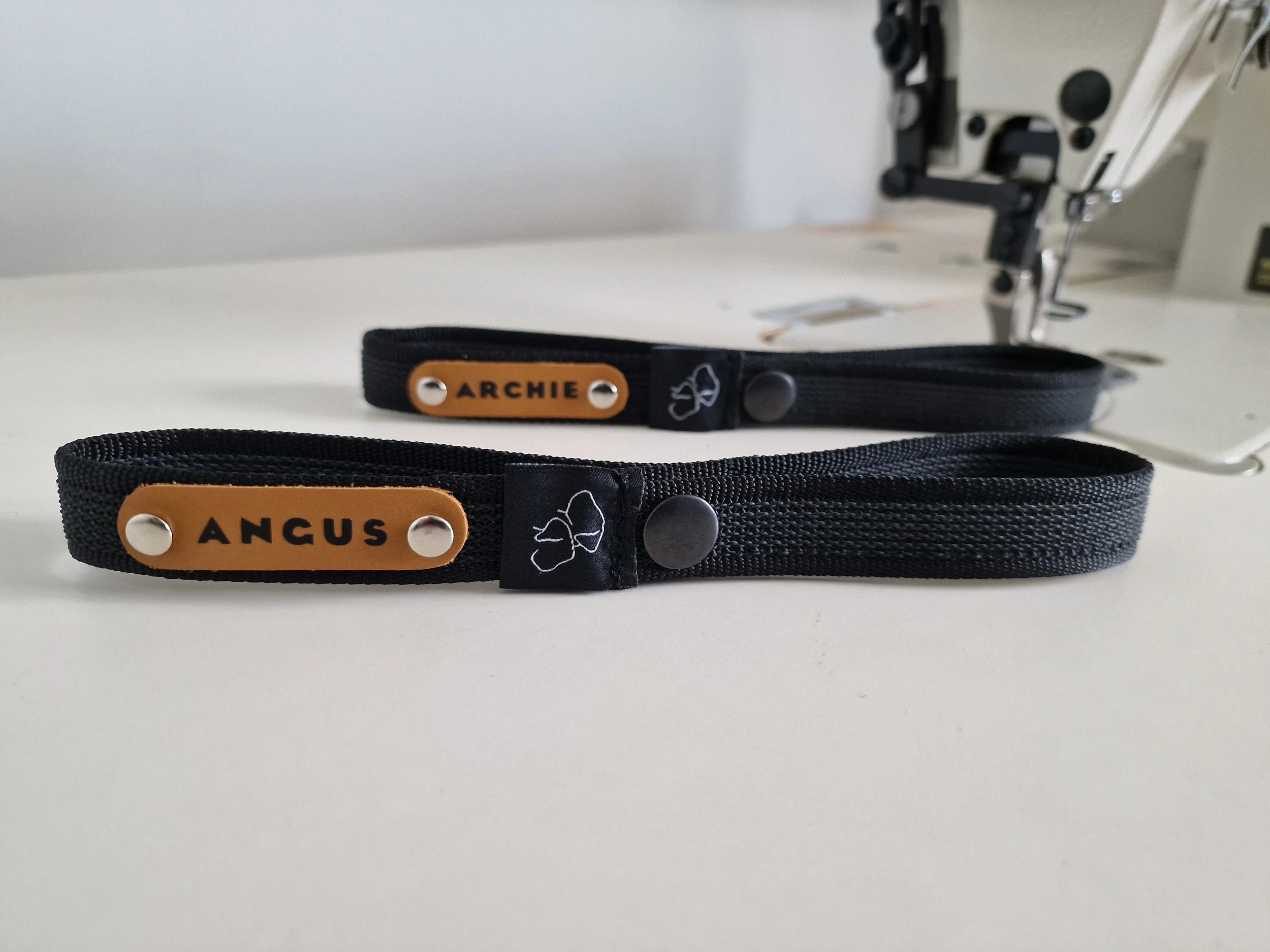 Two black fabric collars with leather accents, one with the name tag 'ANGUS' and an embroidered bow.