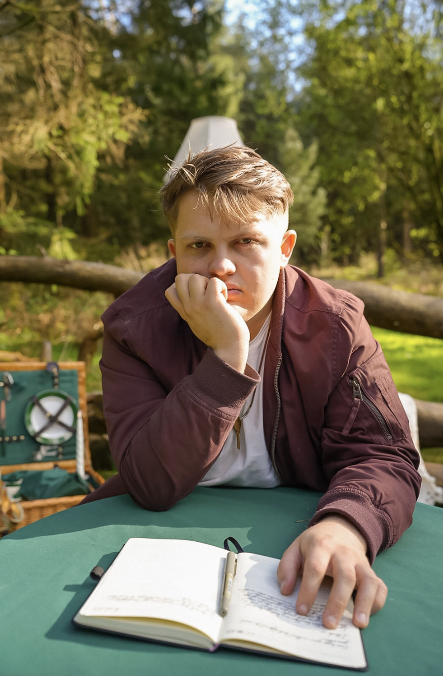 A young man sits at an outdoor table, resting his face on his hand, with an open notebook and a pen in front of him. He is wearing a maroon jacket and has a serious or contemplative expression, with a background of green trees and a picnic basket nearby.