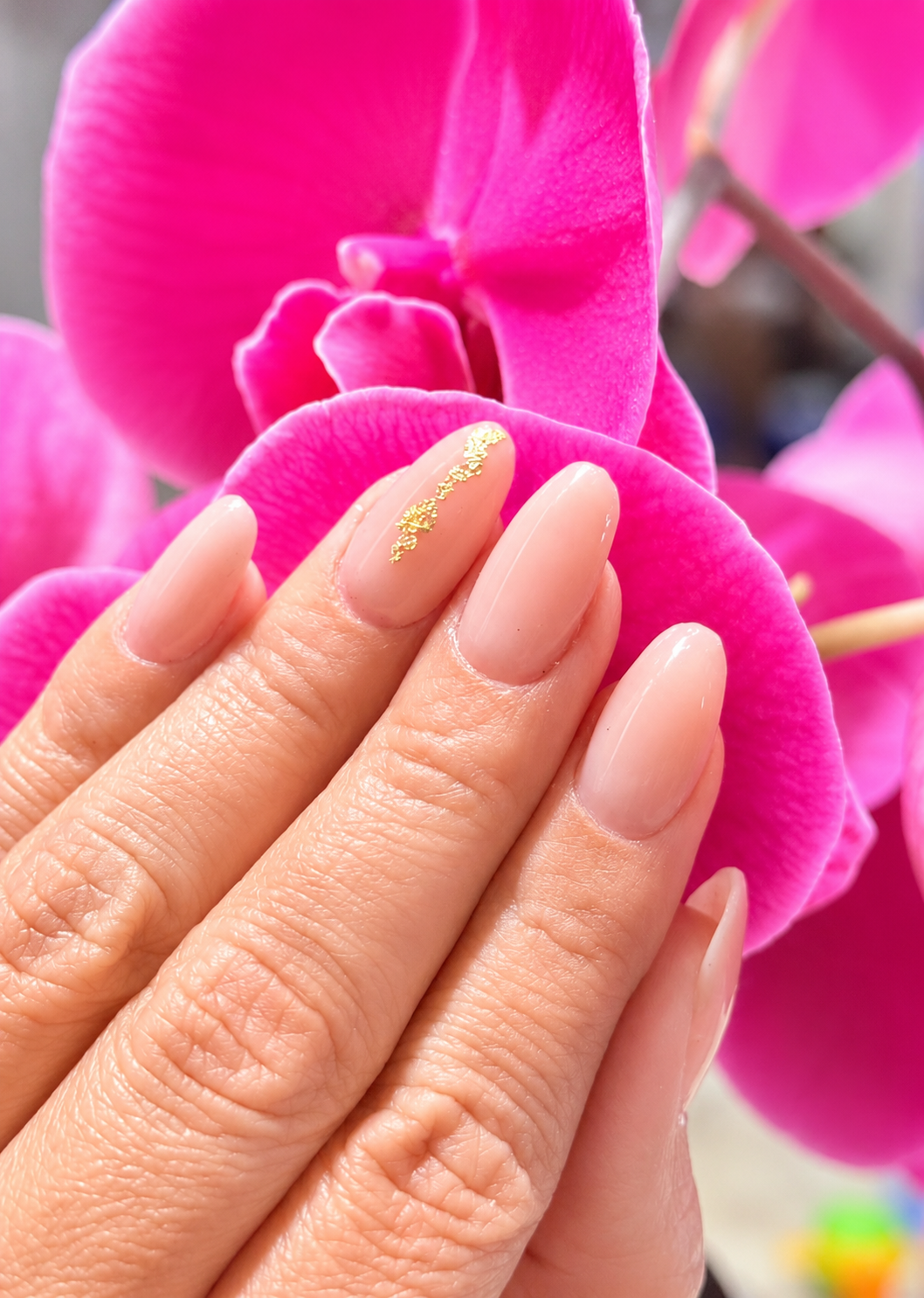 Close-up of a woman's hand with nude-colored, almond-shaped nails with a gold decorative detail on the index finger, resting on vibrant pink orchids.
