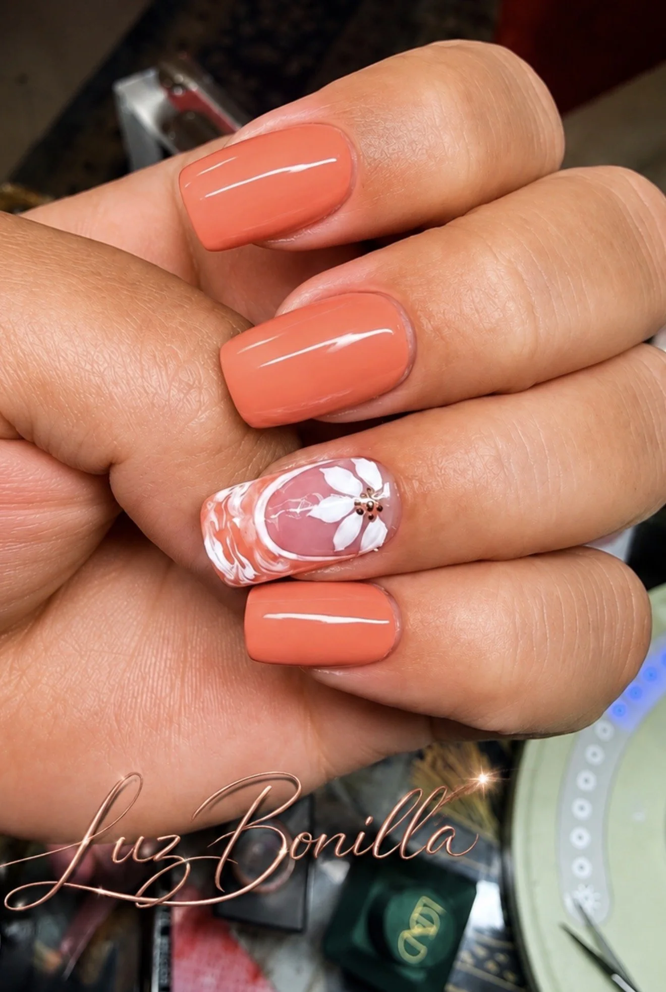 Close-up of manicured fingernails with a coral color, featuring a floral design on the ring finger. The background shows various nail art products.