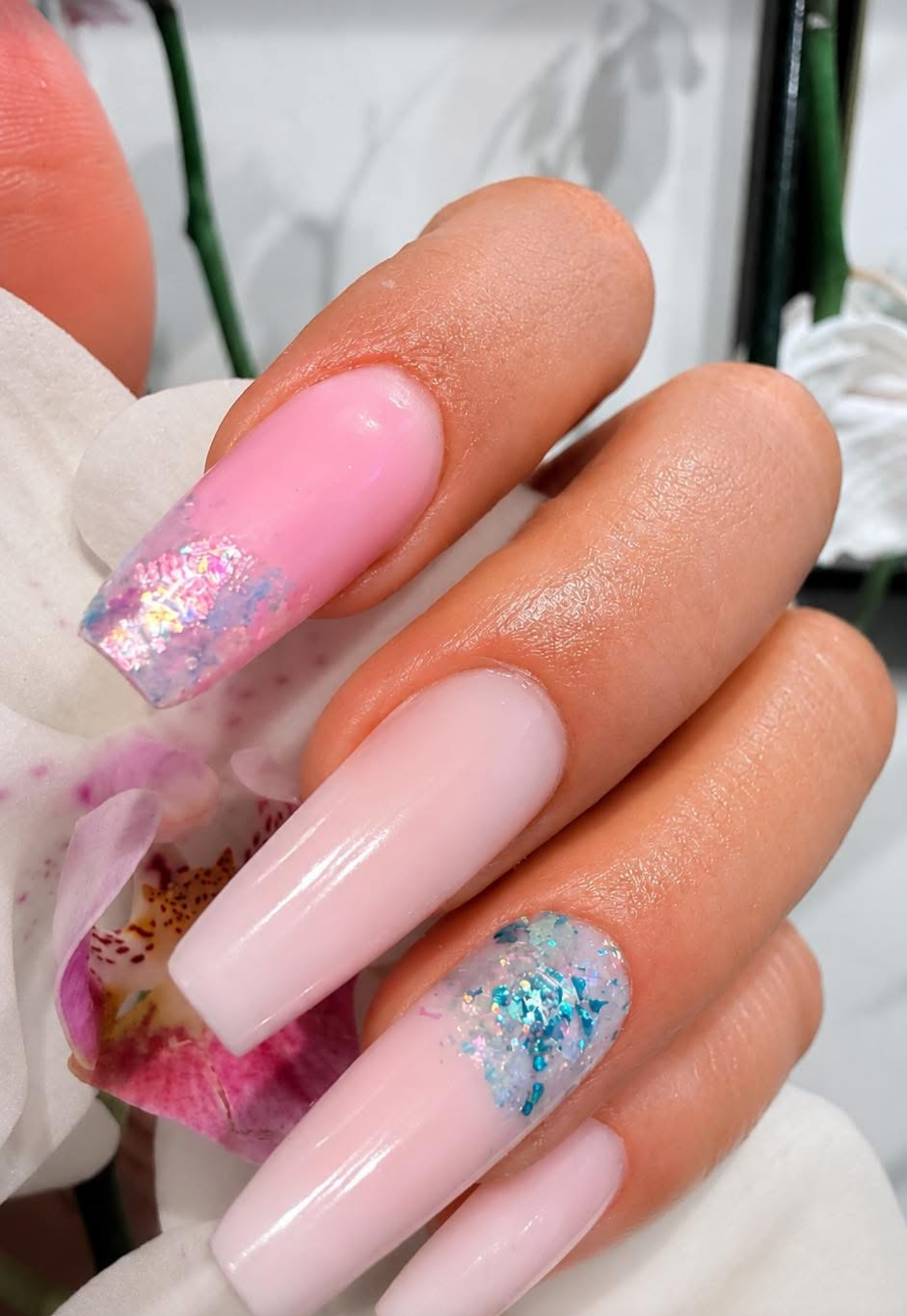 Close-up of a hand with long acrylic nails featuring pastel pink, white, and glitter designs, set against a background with orchids.