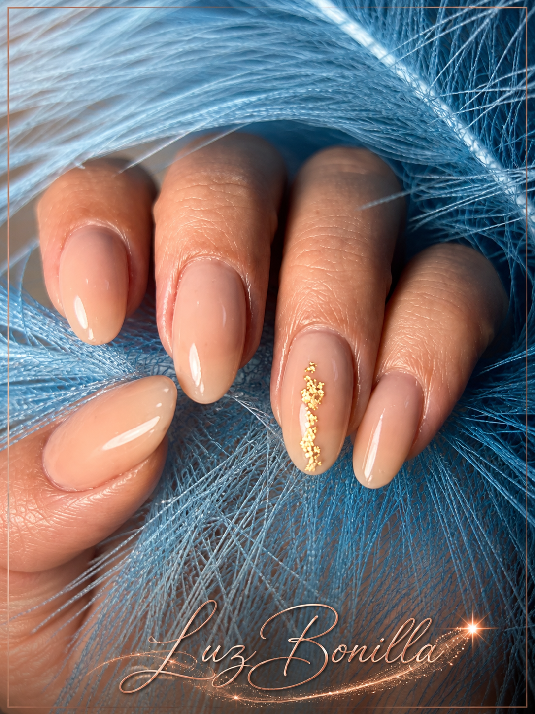 Hand with neatly manicured nails featuring nude polish and gold glitter accent, resting on blue decorative fibers. Text 'Luz Bonilla' at the bottom.
