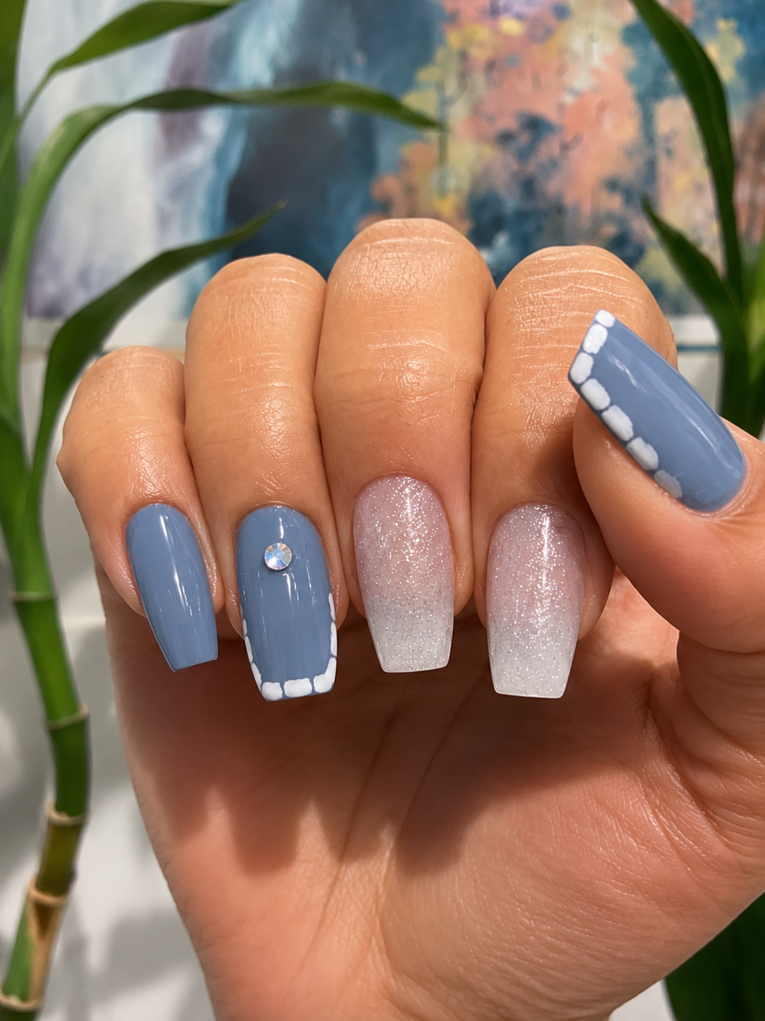 Manicured nails with blue polish, white accents, and a rhinestone, with glittery white and clear designs.