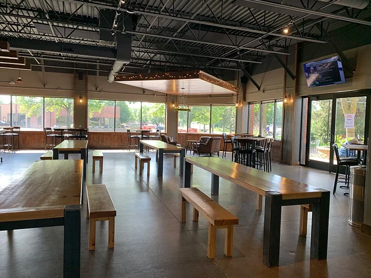 Spacious modern brewery dining room with wooden picnic tables, benches, large windows, and exposed industrial ceilings