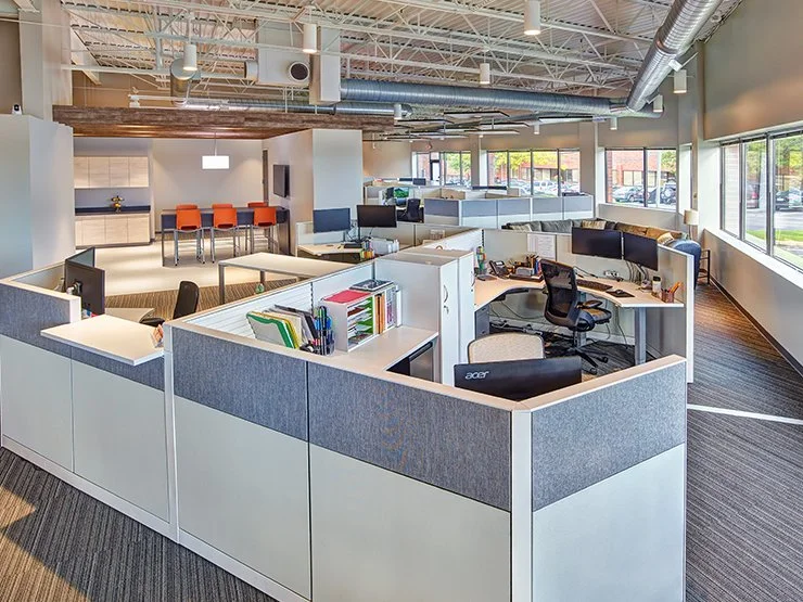 Modern open-plan office with gray cubicles, desks, computers, lounge seating, and a kitchenette