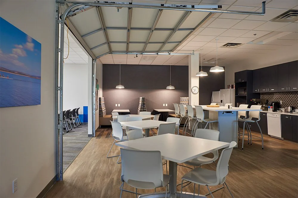 Modern office break room with cafeteria tables, chairs, kitchen, pendant lights, and open garage door