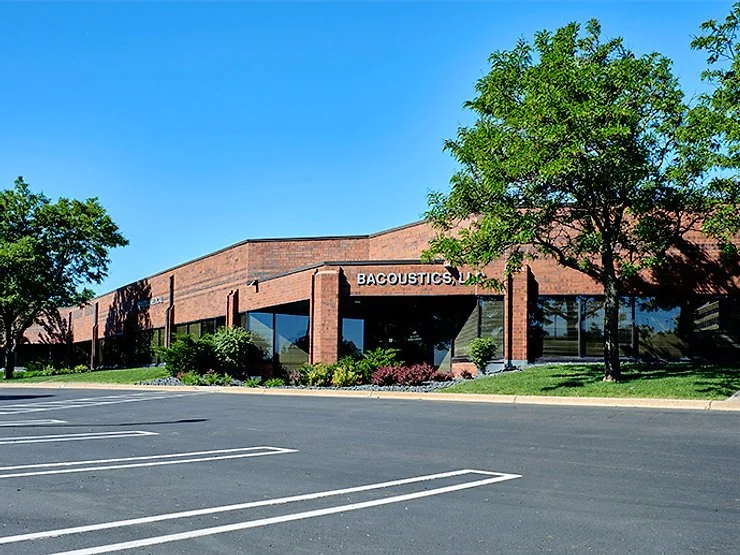 Exterior of a brick commercial building labeled BACOUSTICS, with landscaped grounds and an empty parking lot