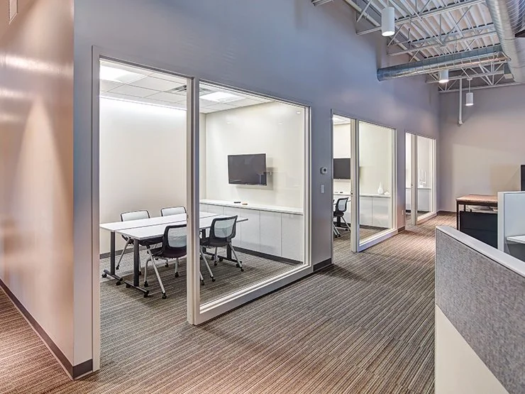 Modern office hallway with glass-walled meeting rooms, conference tables, rolling chairs, and exposed ceilings